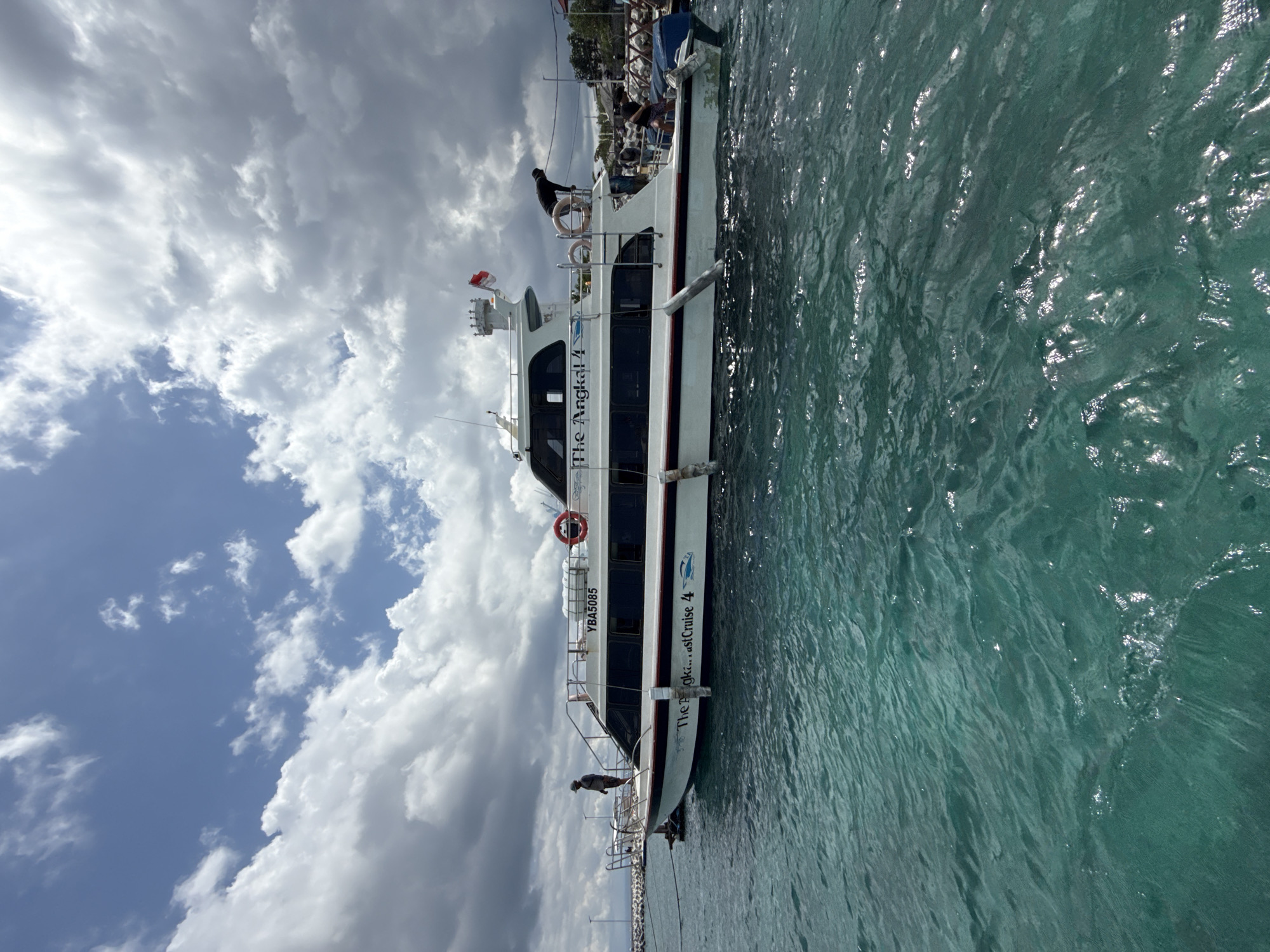 A medium shot captures a boat, labeled "The Angkal..." and "East Cruise 4", docked in the water, with part of a cloudy sky behind it. The water is a mixture of lighter and darker turquoise, with subtle rippling effects.
On the boat, at least two people are visible. One stands on the upper deck, near the ship's controls, while another is towards the front of the boat. Other people are visible, but details are unclear. The boat has a flag at the top and a life preserver attached to its side.
The specific location in Nusa Penida, Indonesia is not ascertainable from the image.
