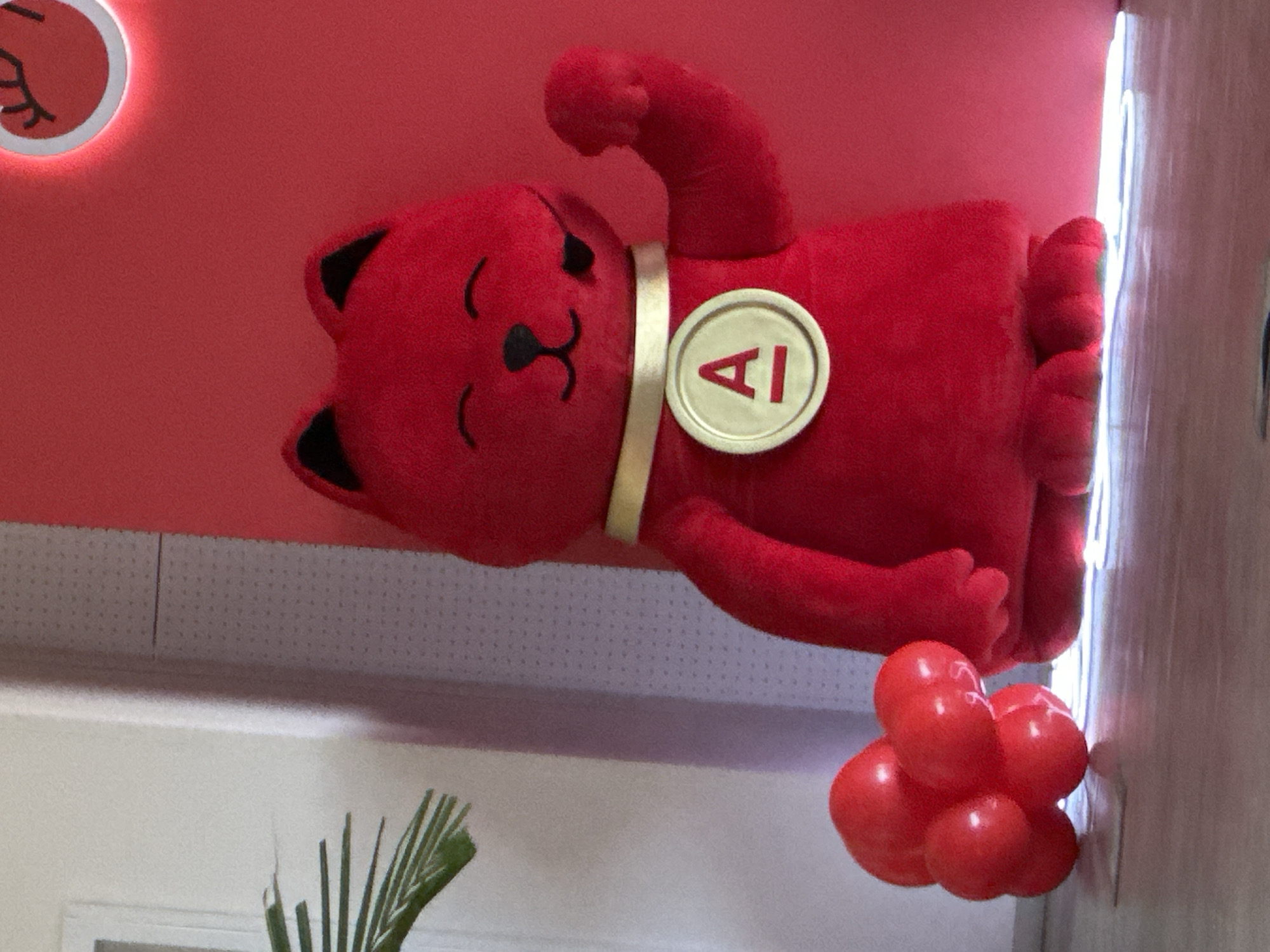 A red plush cartoon animal wearing a collar with a circular 'AI' logo is mounted on a red wall. A small cluster of red balloons hangs below the plush figure. The environment includes a white wall with a palm-like plant in front, and what appears to be an office ceiling. The location is Moscow, Russia.