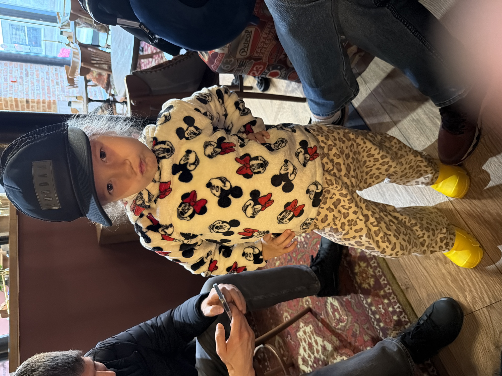 A child wearing a black cap, a white sweatshirt with Mickey Mouse designs, and leopard-print pants with yellow boots is the primary subject. The child is surrounded by adults. The location appears to be indoors, possibly a cafe or restaurant, given the presence of furniture and other people. The location is unspecified and Moscow, Russia cannot be determined from the image. The details suggest a casual, family-oriented setting.
