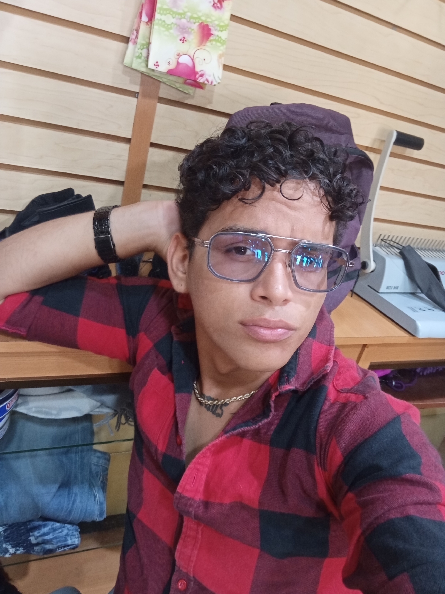 A young man with dark, curly hair and light brown skin is featured in a close-up, eye-level portrait taken in an indoor commercial setting, likely a shop or market stall in Juan Griego, Venezuela. He wears modern, light-tinted rectangular glasses that reflect the ambient light, and a gold-colored chain around his neck. His attire includes an open red and black plaid shirt. A dark-colored watch is visible on his left wrist, which is bent and resting on a wooden shelf or counter.
The background consists of a light-colored wooden slat wall. Colorful fabric items with floral patterns are hanging in the upper left. To the right, behind the man, a light grey machine, possibly a binding machine, bears the text "KZY1 MB". A purple cap is partially visible behind his head. Below the shelf, folded clothes, including blue jeans, are stacked. An object with "OEO" text is also partially visible. The man has a neutral to slightly contemplative expression, looking directly at the camera. The scene is brightly lit, suggesting either daylight indoors or strong artificial lighting.