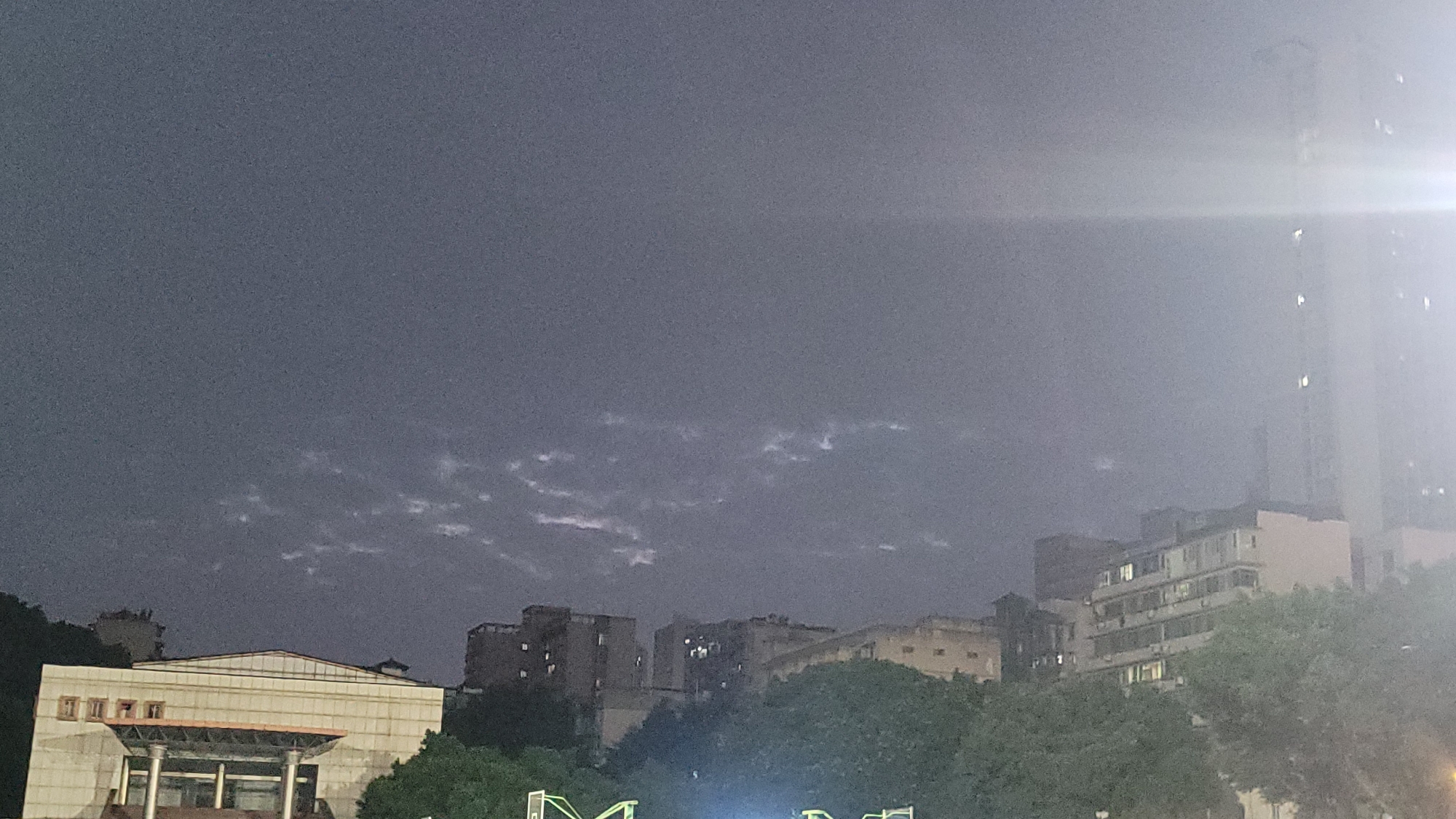 This image captures a quiet urban scene at night or late evening in what appears to be Yuzhong District, China. The sky is dark and largely overcast, with distinct wisps of lighter, stratiform clouds visible across the expanse.
In the lower left, a prominent light-colored building features a sloped roof and a columned entrance, suggesting a public or institutional structure. Moving towards the center and right, various multi-story buildings, likely residential or commercial, are visible, some with illuminated windows indicating human presence. These structures appear nestled amongst hillsides and trees. To the far right, a tall, slender modern high-rise building extends upwards, partially obscured by a very bright light source (possibly a street lamp or building light) which creates a noticeable lens flare. Lush green foliage, indicative of trees or bushes, occupies the lower and mid-ground sections of the image, obscuring the base of some buildings.
No people or specific activities are discernible, contributing to the serene and somewhat atmospheric mood of the night cityscape. The low light conditions contribute to a grainy texture throughout the image. There is no visible text.