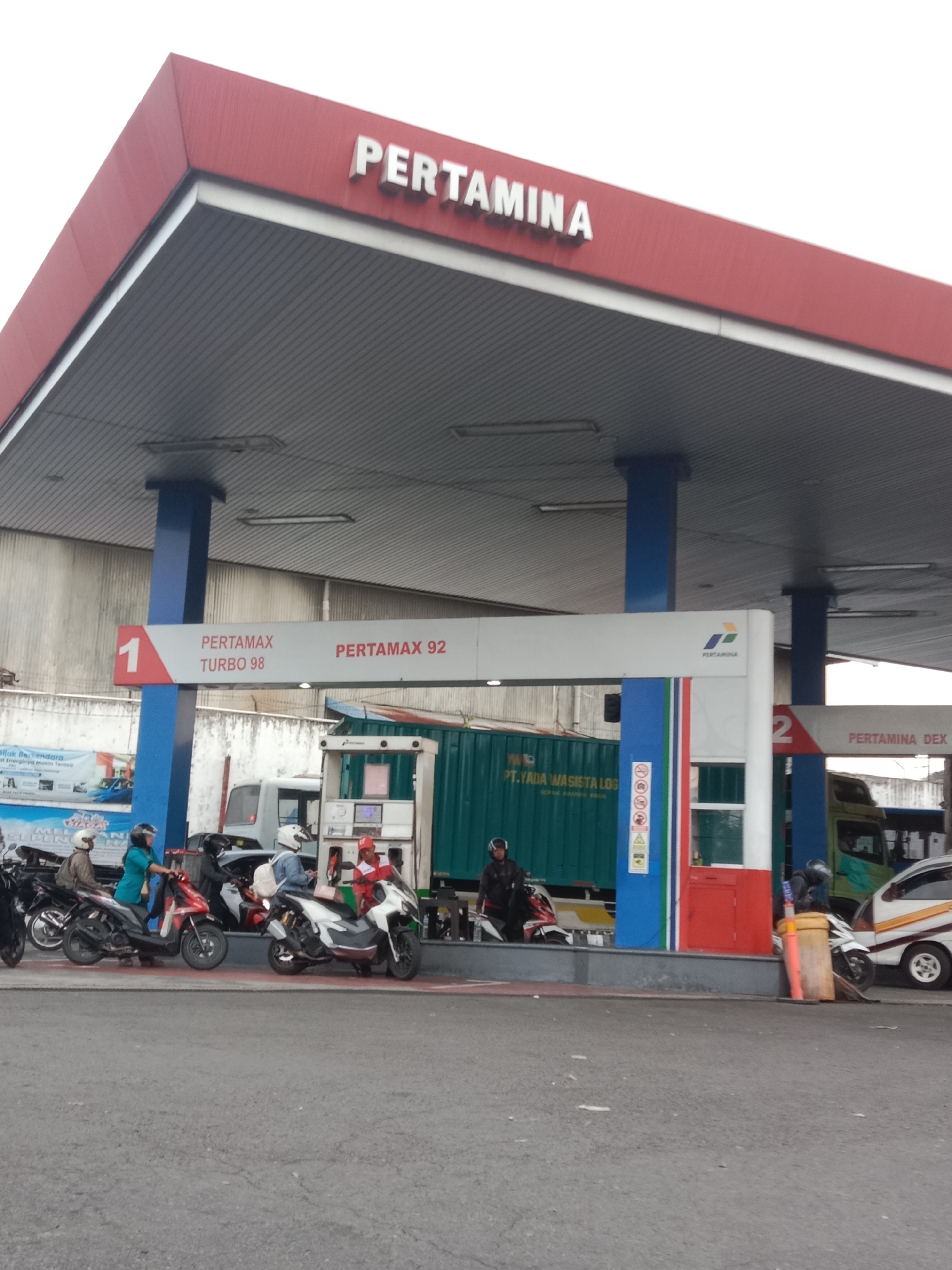 The image depicts a Pertamina gas station, likely located in Semarang, Indonesia, judging by the branding and context. The gas station features a prominent red canopy with the Pertamina logo clearly displayed. Beneath the canopy, several fuel pumps are visible, along with signage indicating different fuel types like "PERTAMAX TURBO 98" and "PERTAMAX 92". A truck with a green container is parked in the background, and a white car with yellow stripes is on the right side of the image. Several people are present, some on motorbikes and one individual in a red uniform who appears to be a gas station attendant. The scene suggests a typical day at a gas station with customers refueling their vehicles. The overall environment is an outdoor commercial space with paved ground.