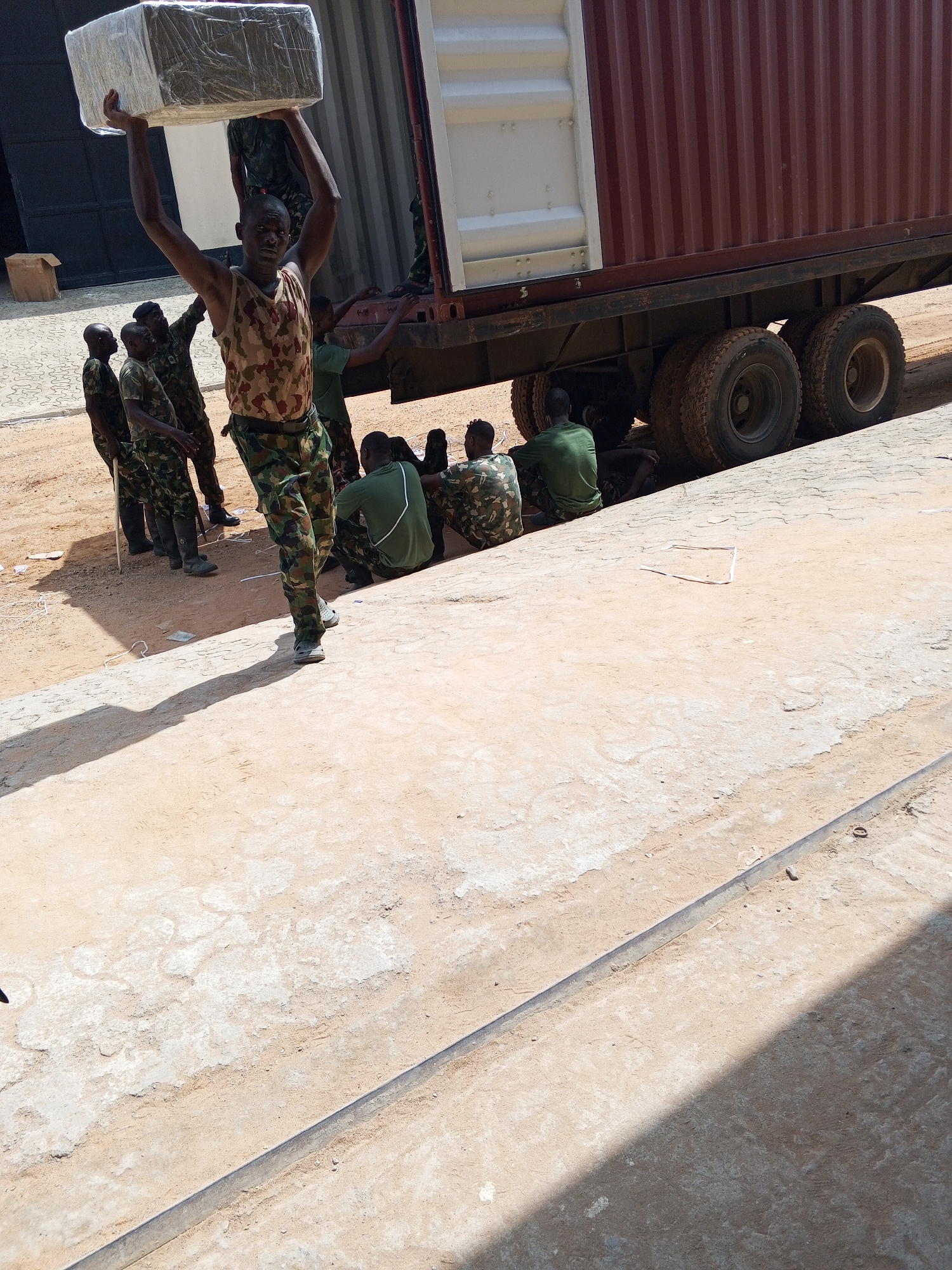 In the city of Unknown, Unknown, a group of people in military-style clothing are gathered near a large truck. One person is carrying a large, wrapped package over their head. Some are sitting or standing near the truck, and others are further back. The ground is sandy and appears to be outdoors.