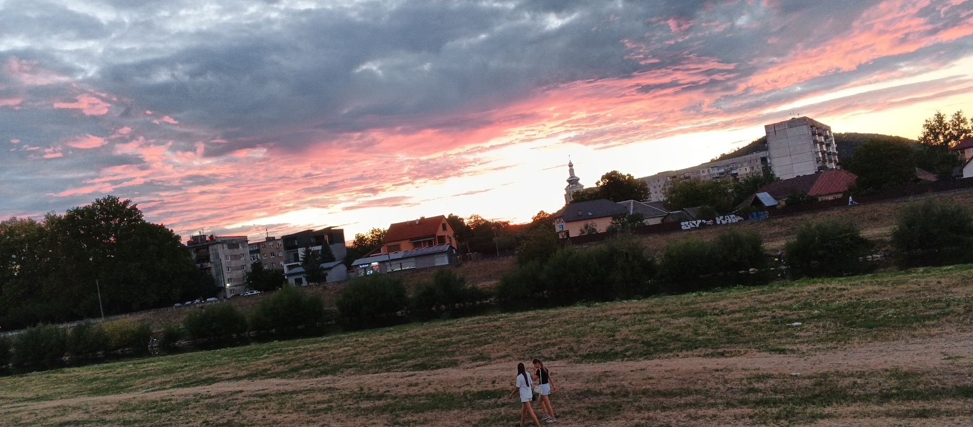 In Mukachevo, Ukraine, a panoramic, high-angle shot captures a twilight scene dominated by a vibrant sky filled with dramatic pink and gray clouds. In the middle ground, a collection of buildings, ranging from residential blocks to smaller houses, are nestled against a hillside. A church steeple is visible among the structures. The foreground features a dry, grassy field with two young women walking from left to right. The overall impression is one of a suburban landscape bathed in the fading light of sunset.