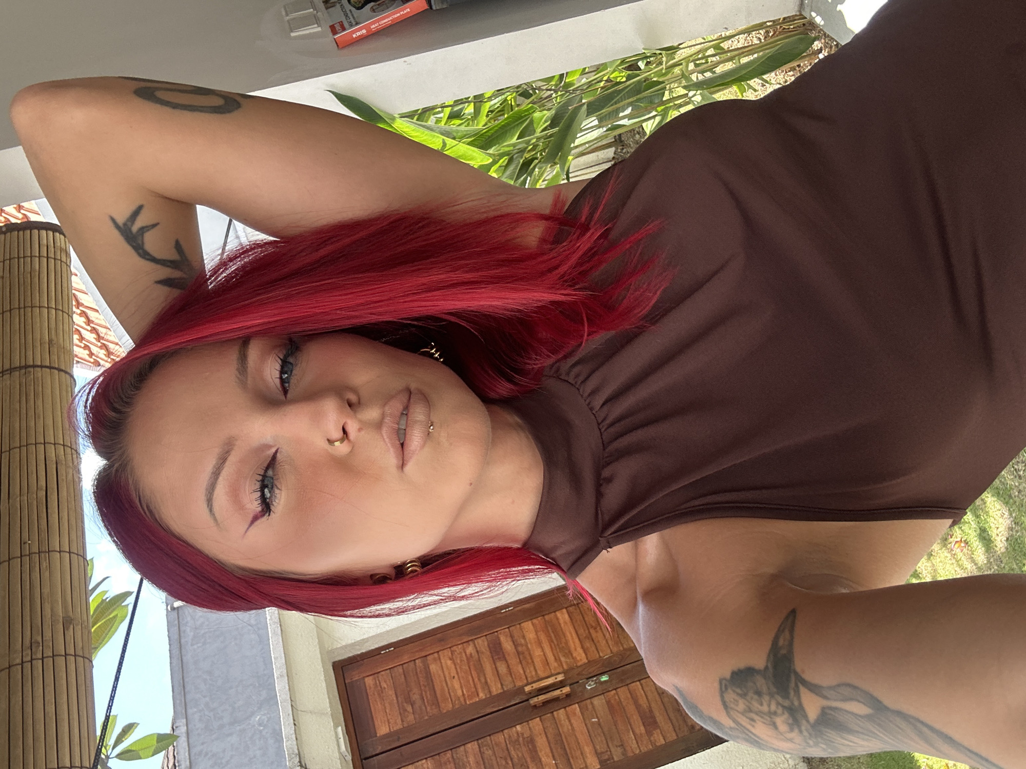A light-skinned woman with red hair is shown in a portrait-style, angled view. She has fair skin, blue eyes, and a small piercing in her left nostril, as well as a piercing above her upper lip. She is wearing a dark brown sleeveless top.
Her right arm is raised above her head, revealing tattoos on her arm and upper body. One tattoo on her forearm is a dark design. There is also a tattoo on her left arm. The background shows a building with a light-colored exterior wall and a section of a bamboo-like structure to the left. A wooden door or panel is visible behind her head, suggesting an indoor or semi-outdoor setting. Outside, there is greenery.
The location is identified as North Kuta, Indonesia.