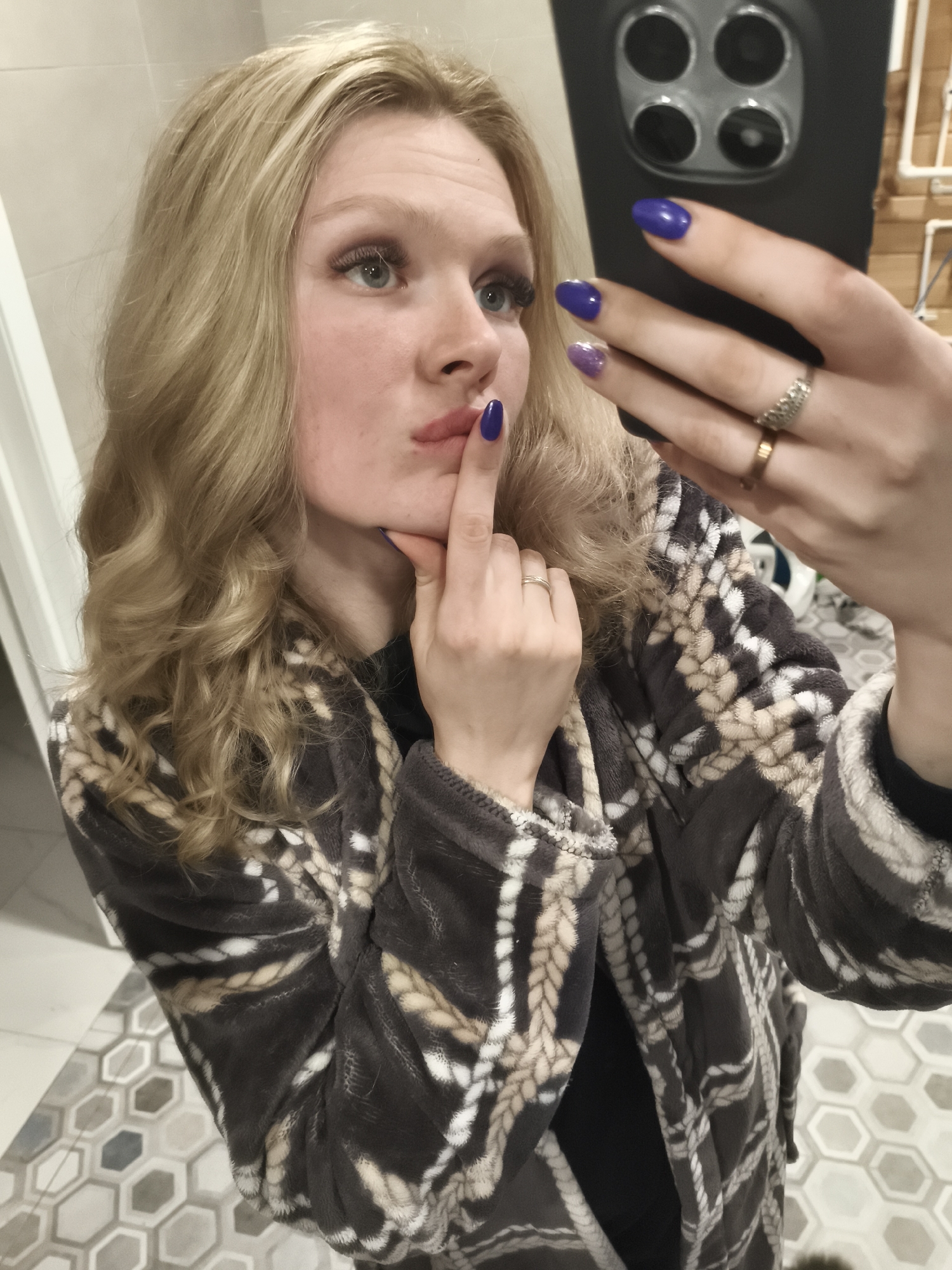 A woman with blond wavy hair and blue nails is taking a selfie with a black smartphone in a bathroom. She is wearing a gray and beige robe and has rings on her fingers. The bathroom has white walls and a tiled floor with a gray and beige geometric pattern. A white fixture is visible in the background. The location is Русский Вожой, Russia.