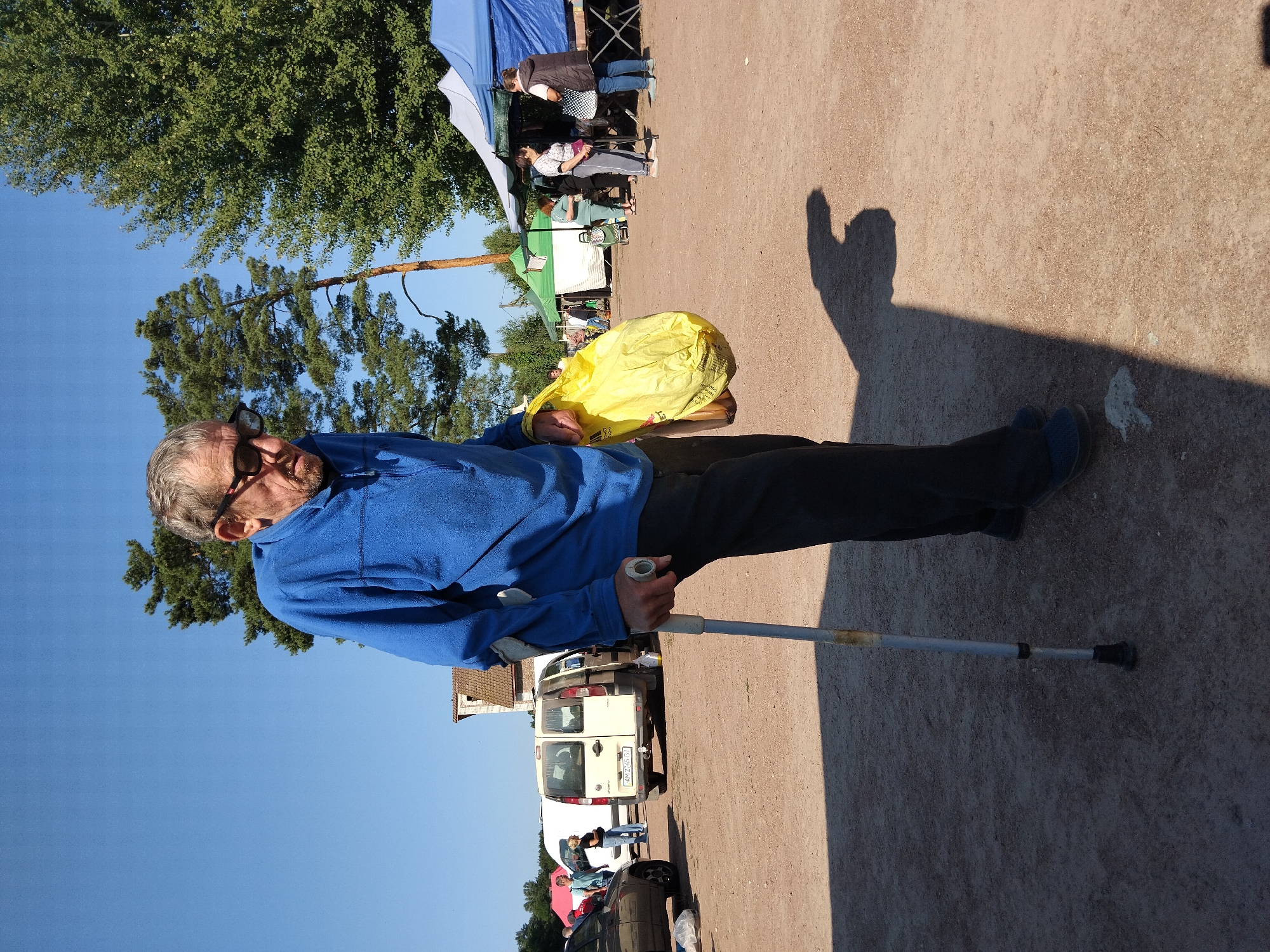 A man, identified by context as being in the city of Irshansk, Ukraine, walks with a crutch and carries a yellow plastic bag. He is wearing a blue fleece jacket and dark pants. Behind him, several tents are set up, suggesting an outdoor market or fair. Vehicles, including vans and cars, are parked nearby, and other people are visible browsing the stalls. The ground is a dirt or gravel surface, and trees and a clear blue sky are in the background. The lighting indicates it is daytime, likely sunny.