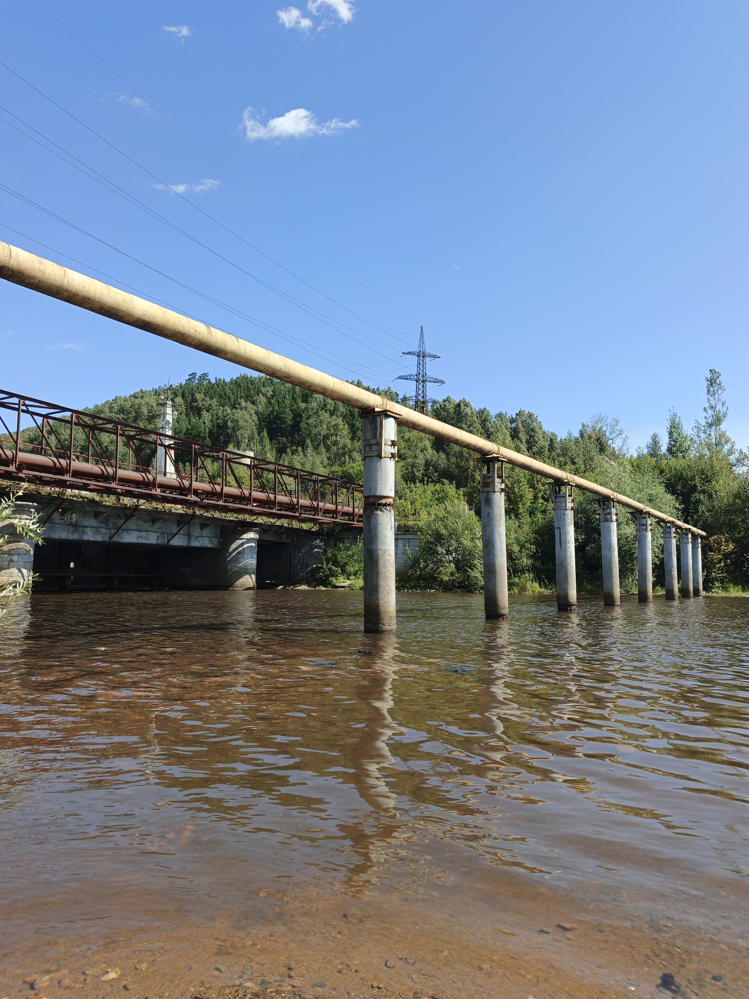 An outdoor, low-angle, wide shot captures a bridge and an overhead pipe crossing a wide, brown river. The sky is a clear, bright blue with a few scattered white clouds. The river's surface is rippled, and the bottom is visible in the foreground, showing sand and small rocks. The bridge, constructed from rusted metal beams and concrete supports, spans the river. Above the bridge, a large yellow pipe, supported by concrete pillars rising from the water, extends horizontally across the frame. On the far bank, a densely wooded hillside rises steeply. In the background, a tall, lattice-like electrical pylon is visible against the sky. The scene appears to be located in Zlatoust, Russia, possibly near the Ufa River or a similar waterway. No people or animals are present in the image.