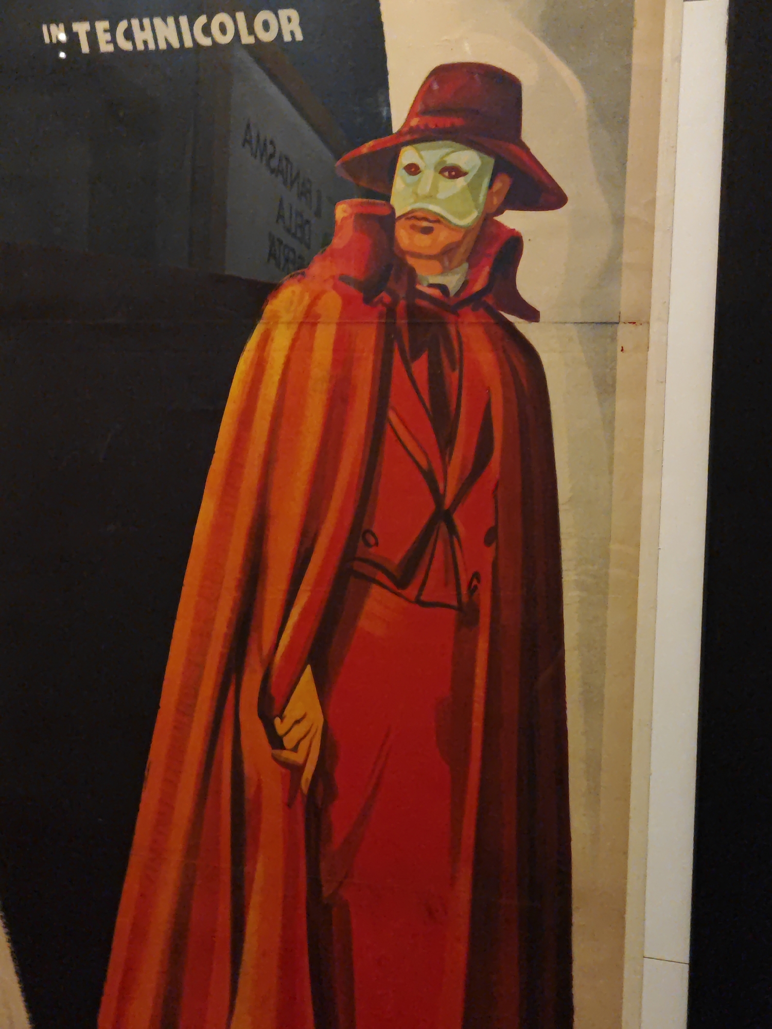 A theatrical poster depicts a figure in a red cloak and a white and green mask, resembling the Phantom of the Opera. The figure wears a red hat and a red suit with black lapels and a black tie. The poster has text at the top that reads "IN TECHNICOLOR." The background is a dark, muted color with some lighter vertical stripes on the right side. There is also some text in a lighter color in the upper left quadrant, some of which appears to be reversed, suggesting it is a reflection or a design element. The poster is framed by a white border on the right and a black border on the left. The poster is displayed in what appears to be an indoor setting, possibly a gallery or a cinema lobby, and given the presence of the poster, it is plausible that this image was taken in Turin, Italy, a city known for its cultural heritage and cinematic history.