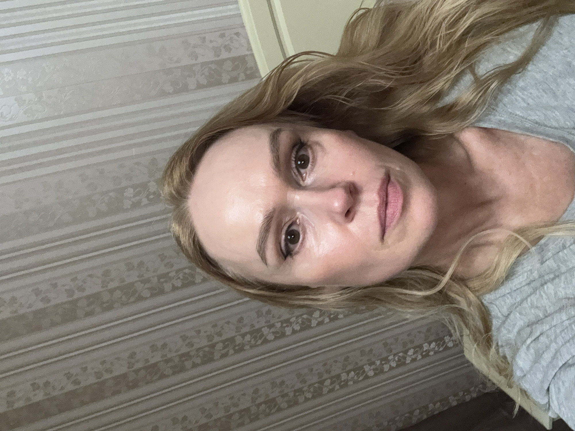 A close-up, slightly tilted selfie shows a woman with light skin, blonde hair, and light eyebrows. She has brown eyes and visible pores. She appears to be wearing a gray shirt. The background consists of striped wallpaper with a floral pattern. The location is likely an interior, possibly a residential building, in the city of Chaikovsky, Russia. There are no visible actions or interactions taking place.