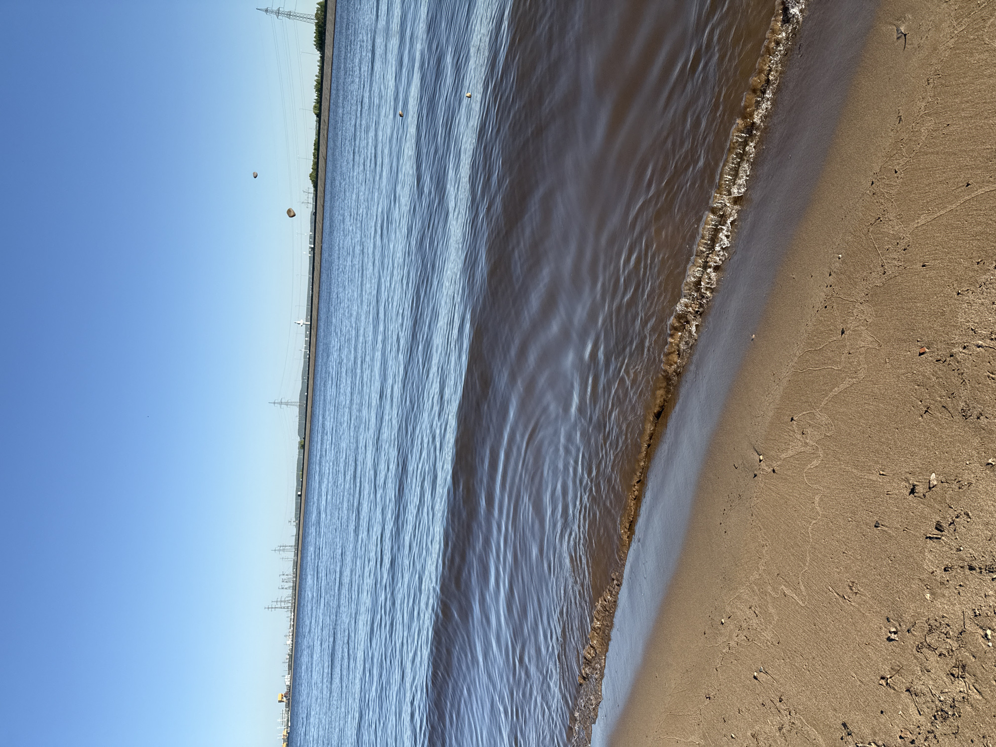 The image depicts a beach scene near Noviy, Russia. The shoreline is visible, with the water meeting the sand. The water is brown and slightly turbulent. The sand is a light brown color. In the background, there are power lines and some vegetation along the shore. Two floating objects are visible in the water. The sky is clear and blue.