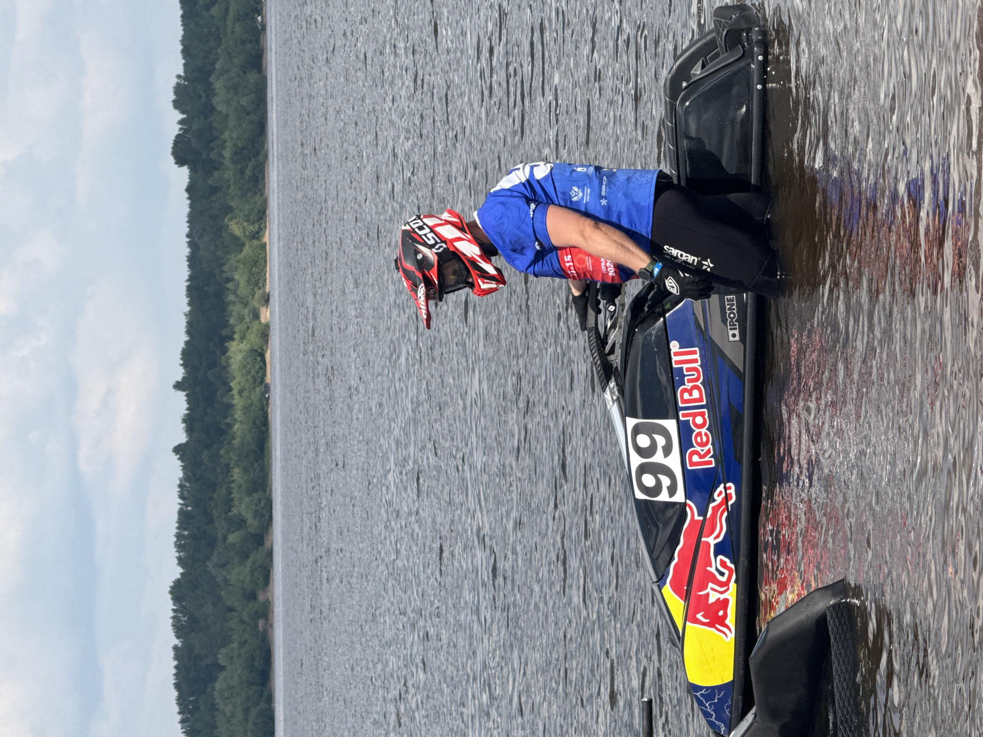 The image shows a person wearing a blue shirt and a red and black helmet, standing next to a jet ski in the water. The jet ski has "99" and "Red Bull" branding on it. The background features a body of water with trees on the horizon. The sky is cloudy. The scene appears to be outdoors. There is no visible indication of the location being in the city of Perm, Russia.