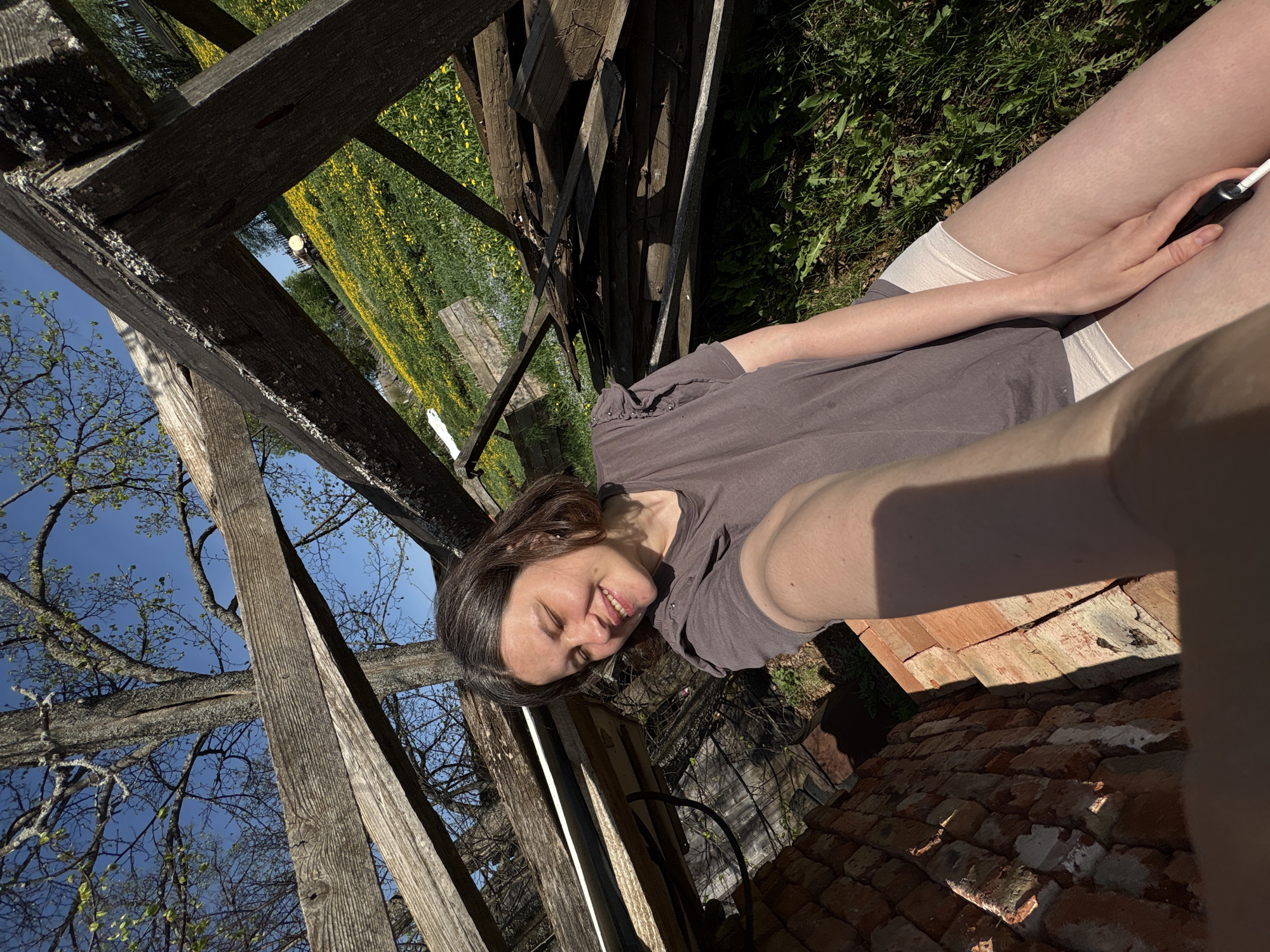 A woman is positioned within a dilapidated structure. The location is Nizhnyaya Chermoda, Russia. The woman is smiling. She is wearing a brown t-shirt and light-colored shorts. She is holding what appears to be a vape pen in her right hand. The structure is made of wood and brick. Beyond the structure is a field of yellow flowers and foliage. Bare tree branches are visible in the background. The sky is blue.