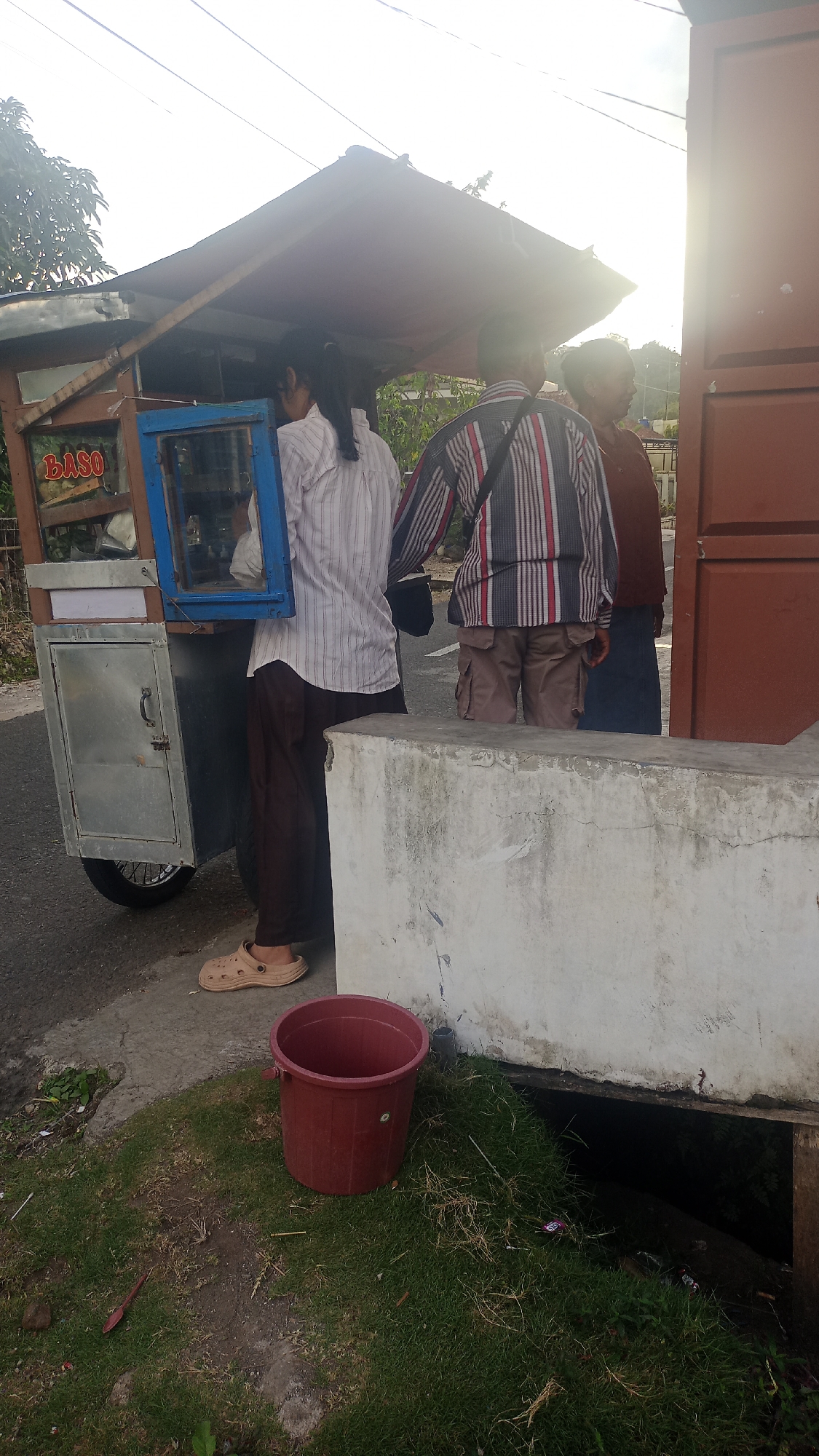 Three individuals stand near a food cart on a street in Pakemitan, Indonesia. The food cart, a mobile establishment with a roof and a glass display case, is labeled "BASO." The person closest to the cart, facing away from the camera, wears a white pinstripe shirt and dark pants. To their right, a second person, also facing away, is dressed in a vertically striped shirt and khaki shorts. A third person, to the right of the second, wears a maroon shirt and dark pants, facing partially towards the camera and smiling. A red plastic bucket sits on the grassy area in the foreground, next to a concrete barrier. The street appears to be paved, and vegetation is visible in the background. The overall lighting suggests it is daytime.