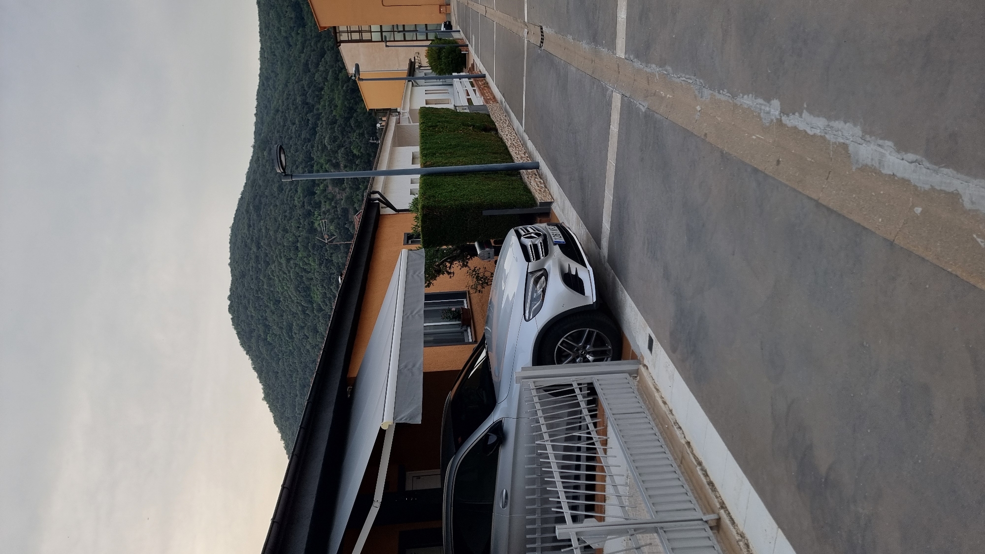 A silver Mercedes-Benz SUV is parked in front of a single-story house with an orange facade and a gray retractable awning. The car is partially covered by the awning. To the right of the house is a neatly trimmed green hedge. Beyond the hedge, a paved walkway leads to other houses and a street lined with lampposts. In the background, a densely wooded hill rises under a cloudy sky. The scene appears to be in a residential area in Sant Joan Les Fonts, Spain. There are no people visible in the image.