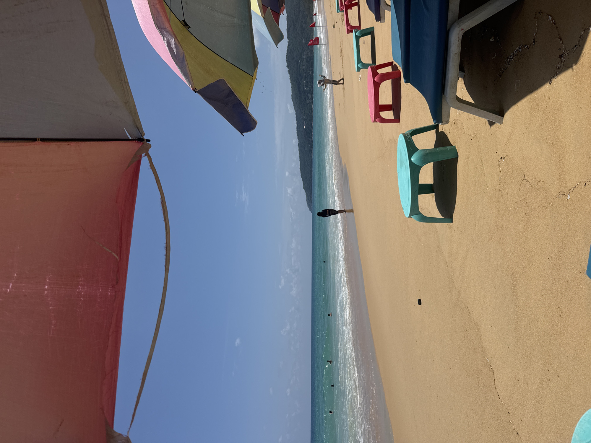 The image depicts a beach scene in Karon, Thailand, under a bright, clear blue sky. In the foreground, a large pink umbrella is partially visible, casting a shadow. Several colorful beach chairs are set up on the golden sand, some with navy blue cushions. Small, brightly colored plastic tables in turquoise and pink are placed in front of some of the chairs.
The midground shows the sandy beach extending towards the turquoise ocean. Small waves are gently breaking on the shore, creating white foam. Several people are visible in the water, swimming or wading. A solitary figure, a woman in a black and white striped dress, stands at the edge of the water, looking out towards the sea. Further down the beach, two more figures, possibly a child and an adult, are standing near some red flags.
In the background, a tree-covered hillside forms the coastline, with a few buildings visible along the shore. The overall impression is a peaceful and sunny day at a popular tourist beach.