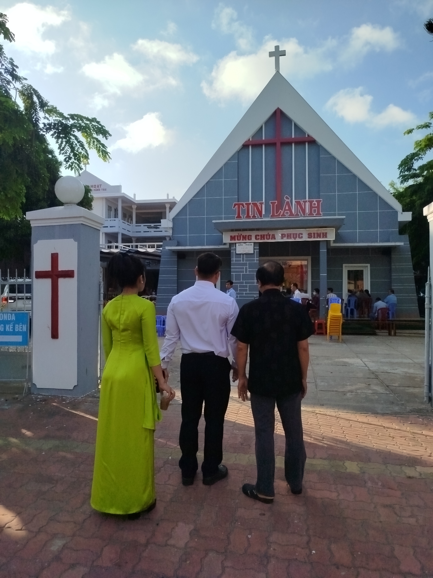Three people from behind walk towards a church in Ho Chi Minh City, Vietnam. The person on the left is wearing a long, bright green dress. The person in the center is wearing a white shirt and black pants. The person on the right is wearing a black shirt and grey pants. The church is light blue with a large red cross on the roof. The Vietnamese words "TIN LÀNH" are visible above the door, with "MỪNG CHÚA PHỤC SINH" beneath. Other people are standing near the entrance to the church. A gate with another red cross is to the left of the church, while other buildings are behind the church. The sky is blue with scattered clouds.