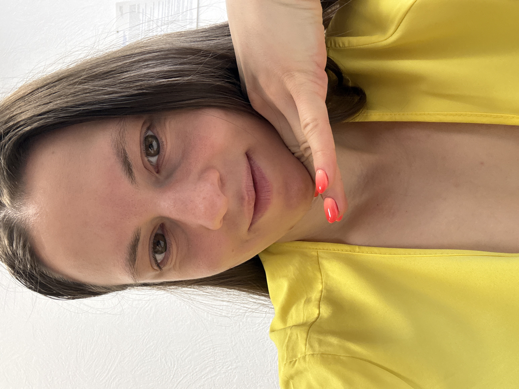 A medium close-up portrait shows a woman in an indoor setting in Chaikovsky, Russia. She has long, dark brown hair that frames her face and falls around her shoulders. Her brown eyes are open and gaze directly at the viewer, and her expression is soft and neutral, with a faint, gentle smile playing on her lips. Her complexion appears natural, with some slight redness visible on her cheeks and forehead.
She is dressed in a bright, vivid yellow garment, possibly a satin or silk shirt, which can be seen on the right side of the frame, showing its lustrous texture. Her right hand is raised and gently rests against the side of her face and neck, partially obscuring her jawline. The fingernails on this visible hand are painted in a distinct ombre style, transitioning from a lighter orange shade at the base to a vibrant red at the tips.
The background consists of a plain white wall, which exhibits a subtle textured pattern. In the upper part of the image, a blurry, light-colored structure, possibly a window frame or another architectural element, is faintly discernible. The scene is illuminated by soft, even lighting, suggesting a bright daytime environment. There is no visible text in the image.