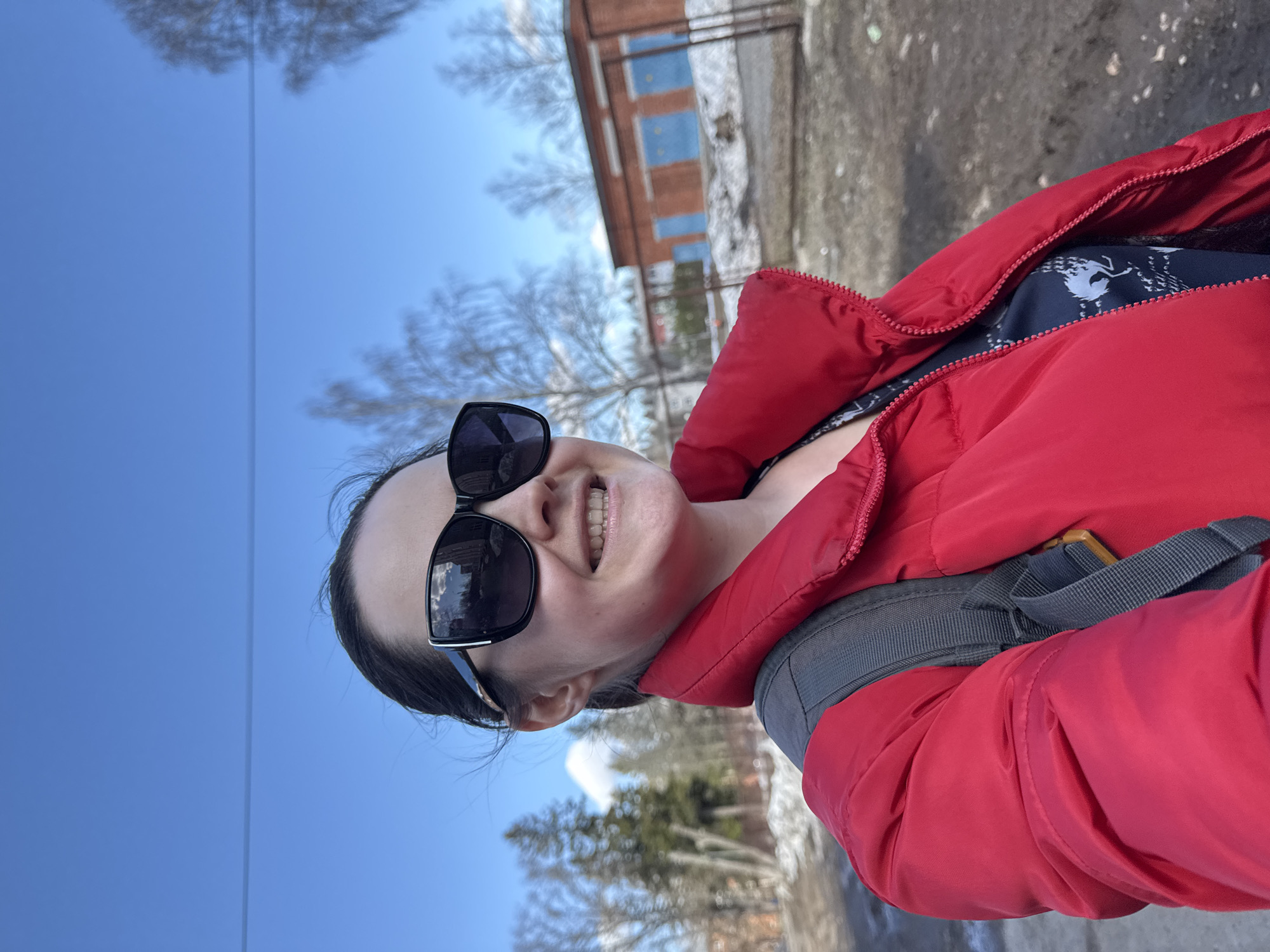 A woman wearing a red puffer jacket and sunglasses is taking a selfie outdoors. She is smiling, and a backpack strap is visible over her jacket. The background includes a clear blue sky, bare trees, a brick building with blue windows, and patches of snow on the ground. The location is likely Izhevsk, Russia.