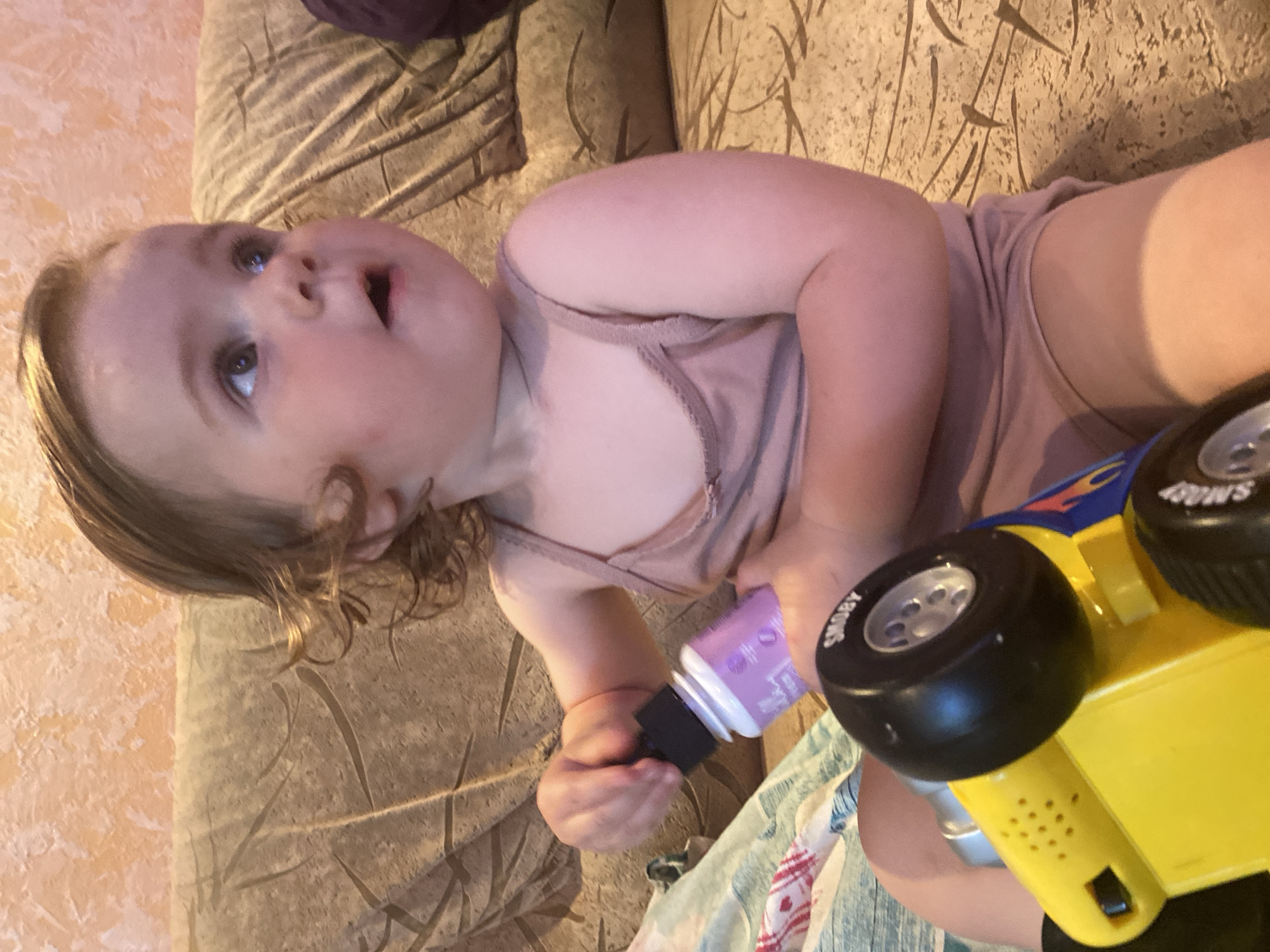 A young child is sitting on a sofa and holding a toy car. The child is wearing a light pink tank top and shorts. The toy car is yellow with blue and red flames. The sofa is upholstered in a textured fabric that appears to be a light brown or beige color. The background wall has a textured, peach-colored finish. The lighting in the scene is warm, suggesting indoor ambient light.