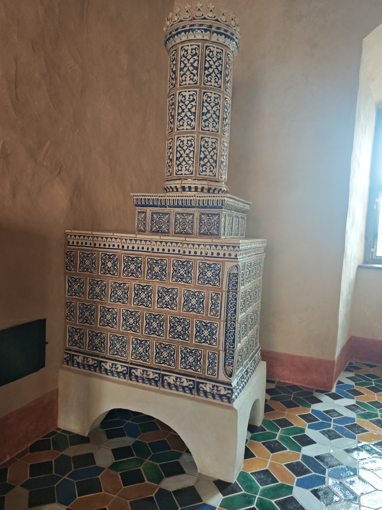 A tiled stove stands in a room with a patterned floor. The stove is tall and ornate, with blue and white tiles covering its surface. The tiles have a decorative pattern of swirls and floral motifs. The stove is supported by a light-colored base with an arched opening. The floor is covered in a geometric mosaic of colorful tiles, including shades of blue, green, yellow, and brown. The walls of the room are a neutral beige color. A window is visible on the right side of the image, letting in a bright light.