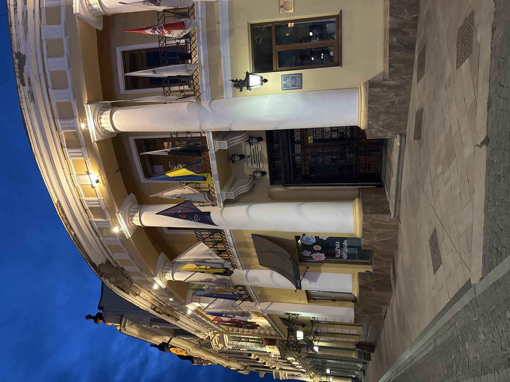 The image captures the facade of a building in Chernivtsi, Ukraine, at dusk. The building is light yellow with classical architectural elements, including large white columns and ornate cornices. Several flags are displayed on balconies along the upper level, including what appear to be Ukrainian national flags. The street in front of the building is paved with cobblestones and light-colored tiles, and there are several lampposts illuminating the area. The sky is a deep blue, indicating the transition from day to night. There are no people visible in the scene. A sign on one of the windows reads "ПОДАРУНКИ НАЙРІДНІШИМ" which translates to "Gifts for the dearest".