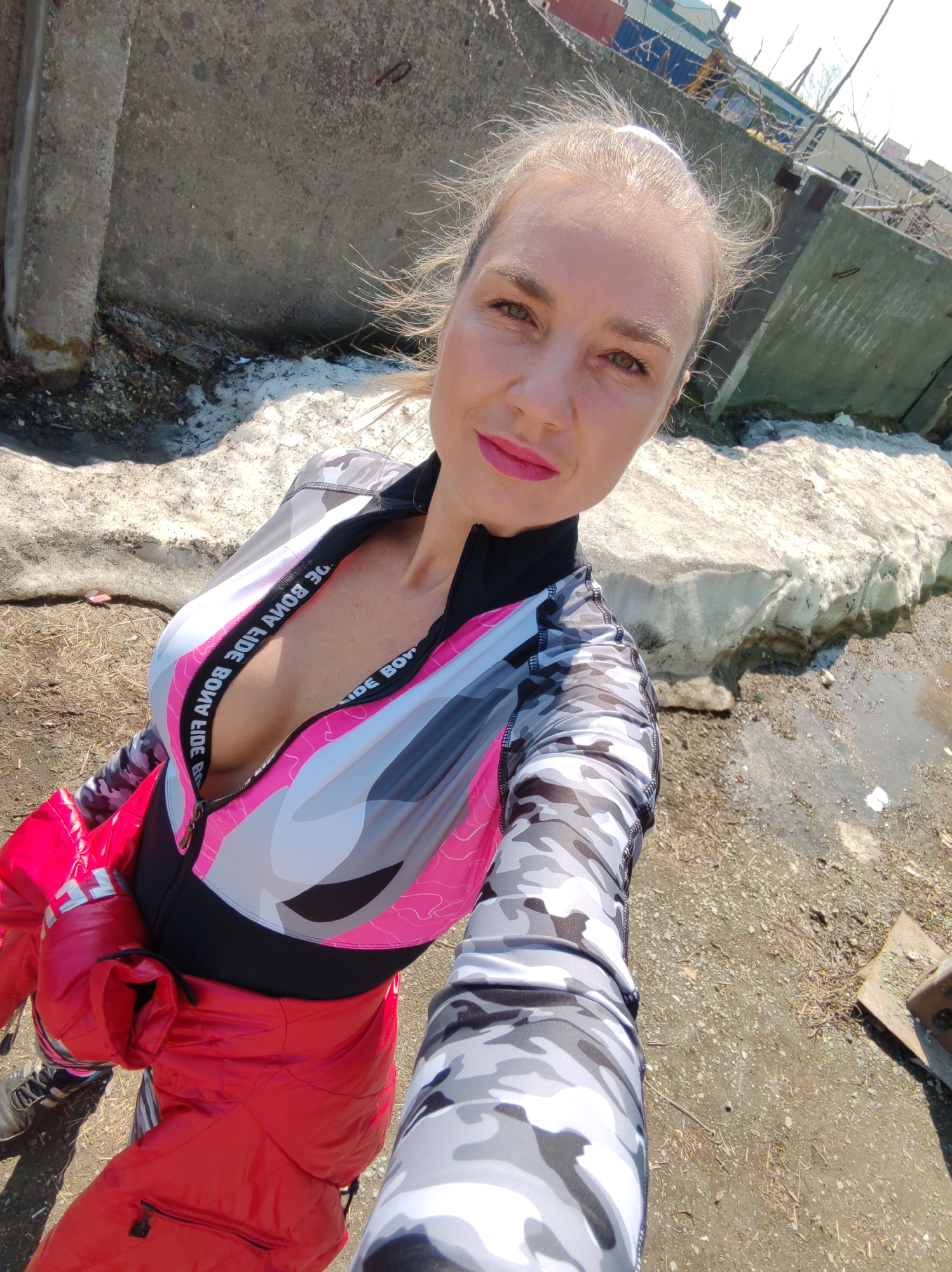 A blonde-haired woman, with her hair pulled back and wisps blowing in the wind, is captured in a close-up selfie in an outdoor environment in Petropavlovsk-Kamchatsky, Russia. She looks directly at the camera with a slight smile, wearing pink lipstick and sporting green-brown eyes. She is dressed in an athletic, long-sleeved top featuring a grey, black, pink, and white camouflage pattern on the torso and solid grey/white camouflage on the sleeves. The top is partially unzipped, revealing the inner trim with the text "BONA FIDE" and her cleavage. A bright red puffy jacket is either tied around her waist or draped over her lower body. The lower part of grey/white camouflage leggings is visible on her left leg, along with a portion of a sneaker. The setting appears to be an urban outdoor space with visible signs of a cold or transitional season. The ground is a mix of dirt, gravel, and sparse dry vegetation. To the right, a large irregular mass of melting snow and ice lies next to a muddy puddle. In the background, there are various concrete structures: a dark, weathered concrete pillar on the left, and large, grey concrete slabs or walls appearing somewhat dilapidated or part of an industrial complex, with thin branches or rebar protruding. Further back, obscured industrial buildings with blue and white elements are partially visible under a bright sky. The scene is brightly lit by natural sunlight.
