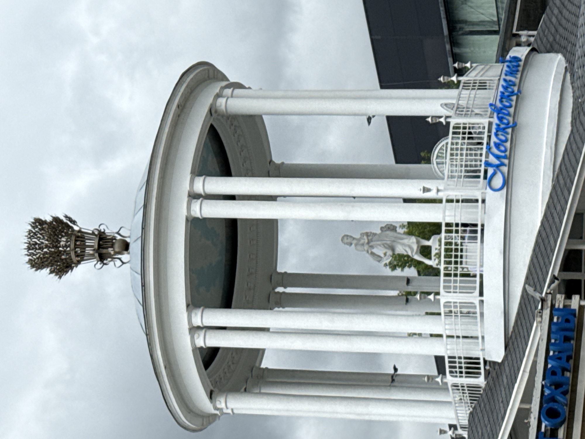 The image shows a white, multi-tiered rotunda structure with a domed roof topped by a wheat sheaf ornament. The rotunda is supported by numerous white columns and features a white railing along its upper level. Inside the rotunda, a white statue of a woman holding a child is visible. The structure is situated in an outdoor urban environment, with a dark building visible in the background. Blue lettering that reads "Москвоское море" (Moskovskoye More, meaning Moscow Sea) is present on the lower section of the rotunda. Another sign with blue lettering, partially visible at the bottom of the image, reads "ОХРАНИ" (Ohrani, meaning Guard or Protection). The sky is overcast and gray. The overall scene suggests a monument or a decorative structure in a public space in Moscow, Russia.