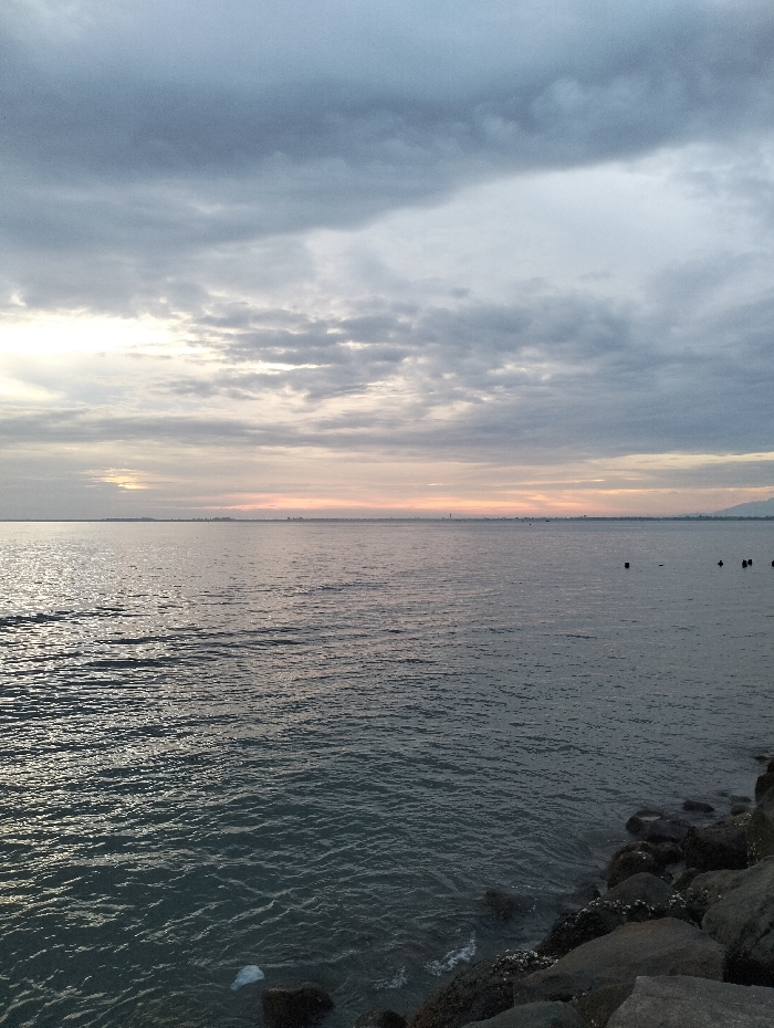 A seascape under a cloudy sky. The sea is calm with small waves reflecting the light. In the distance, a faint skyline is visible. Dark rocks line the shore in the foreground. The sky is filled with grey clouds, hinting at either a sunrise or sunset, with a pale orange glow on the horizon.