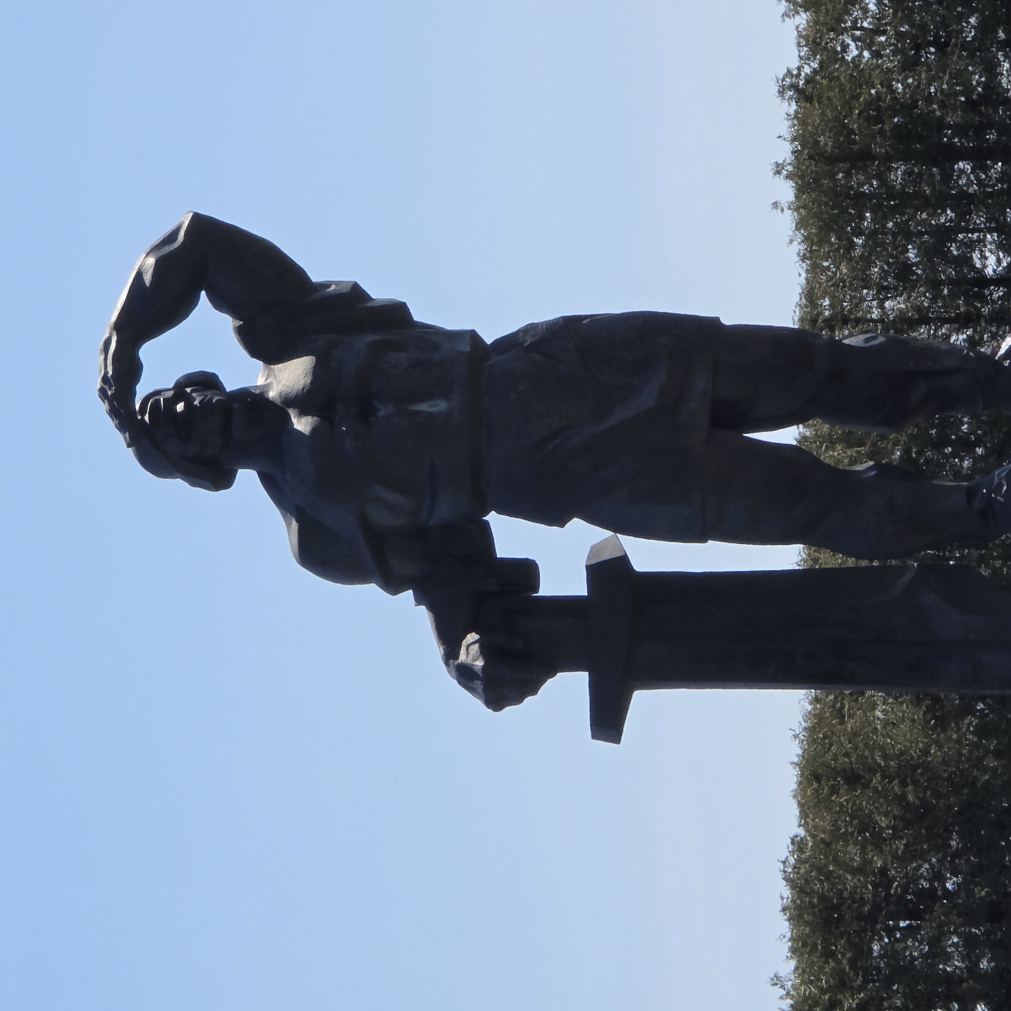A statue of a man stands outdoors against a blue sky and the silhouette of trees. The man is muscular and appears to be holding a large object, possibly a hammer or a sword. He is looking down and his expression is serious. The photo was taken in Свіслацкі сельскі Савет, Belarus.