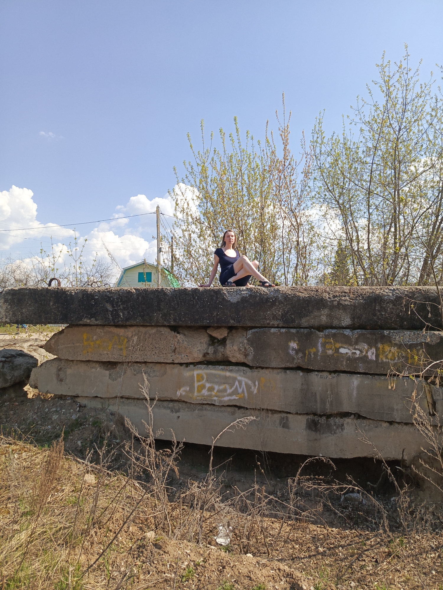 The image depicts a woman sitting atop a stack of large, weathered concrete slabs in a location that may be the city of Chaikovsky, Russia, based on visual clues.
The woman is fair-skinned, with long, light-colored hair, wearing a dark, knee-length dress and sandals. She is sitting with her legs crossed.
The concrete slabs are stacked horizontally, with visible graffiti and discoloration. Behind the stack of concrete are sapling trees and a house with a green roof, which is situated behind a power pole.
In the foreground is dry vegetation and dirt. The sky is blue with scattered clouds. It is daytime, and the lighting suggests a sunny day.