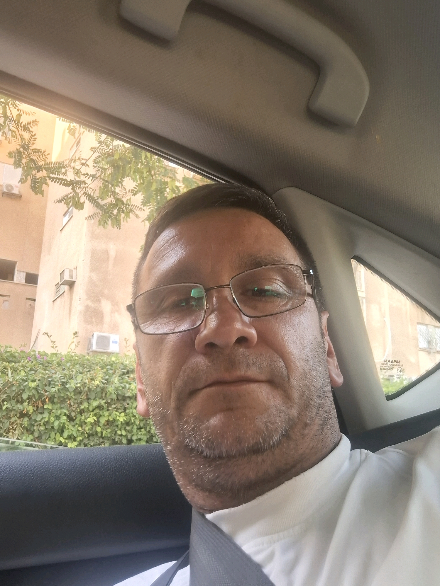 A man is taking a selfie inside a vehicle. He is wearing glasses and a white t-shirt. The background shows a building with several air conditioning units, green foliage, and a window with text that appears to be a car's rear windshield sticker. The building and foliage are consistent with the environment of Be'er-Sheva, Israel.