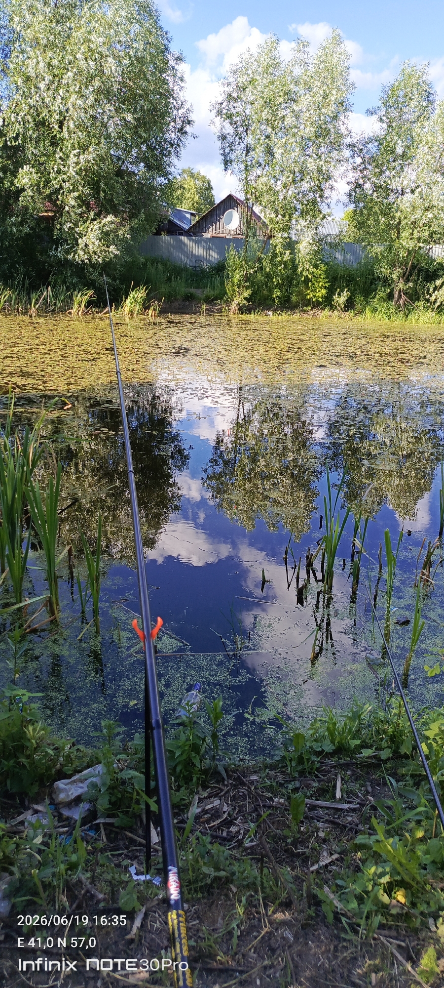 The image depicts a serene natural scene with a body of water covered in lily pads. The water reflects the bright blue sky with scattered clouds and the surrounding trees and vegetation, creating a mirror-like effect. Two fishing rods are visible in the foreground, suggesting a leisurely fishing activity. The vegetation along the bank is lush and green, with tall reeds growing in the water. In the background, a wooden structure resembling a house with a circular window is partially visible, surrounded by trees and a fence. The overall atmosphere is peaceful and tranquil, capturing a moment of nature and relaxation. The timestamp at the bottom of the image indicates the photo was taken on 2026/06/19 at 16:35, with coordinates E 41.0 N 57.0. The device used for the photo is an Infinix NOTE 30 Pro.