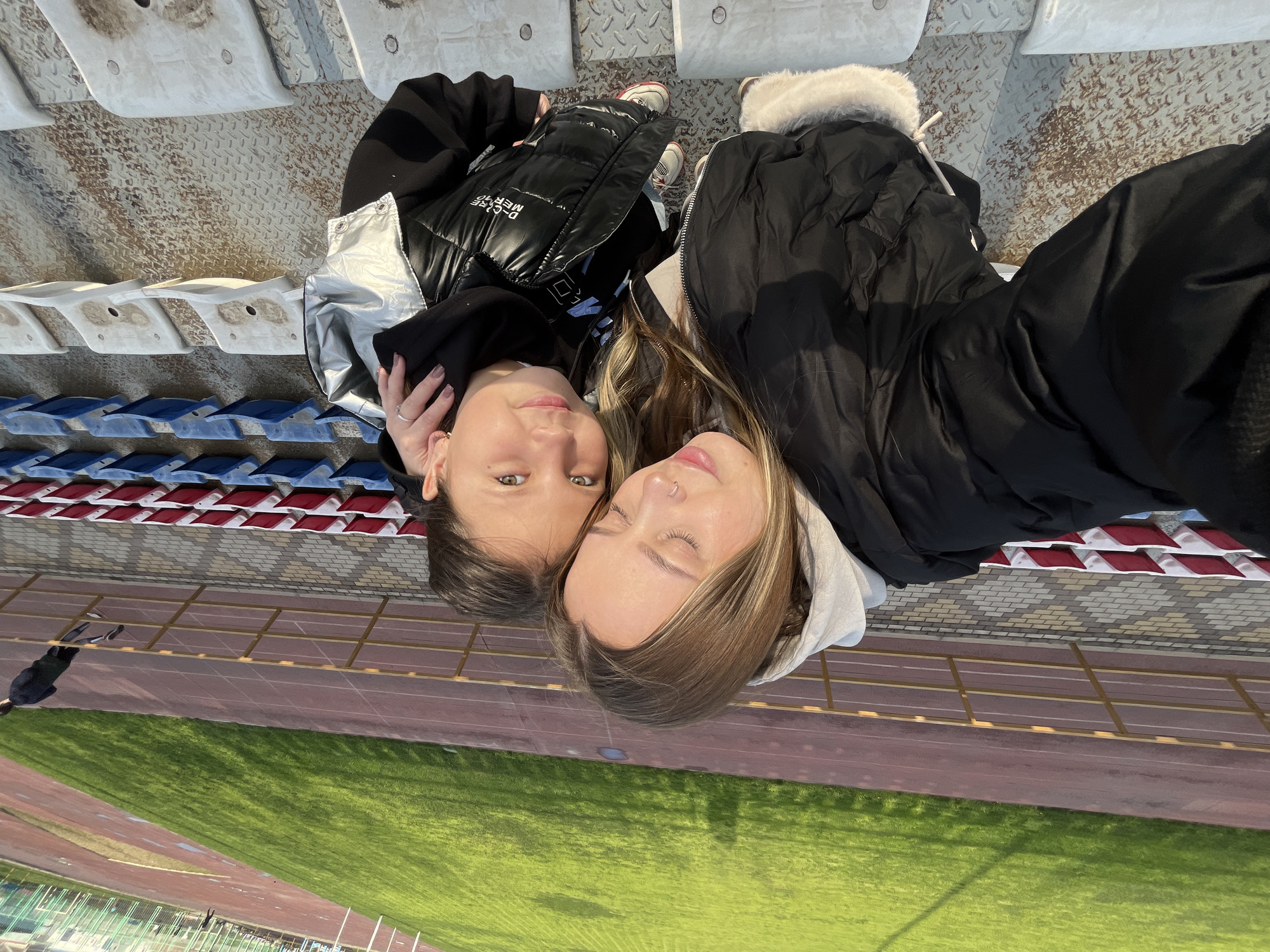 Two young women are lying down at what appears to be a stadium. They are positioned head to head, facing the camera. The woman on the left has dark hair and is wearing a black and silver puffer vest over a black hoodie. The woman on the right has blond hair and is wearing a black jacket with a fur-trimmed hood. The stadium features seating and a running track next to a grass field. The image appears to be taken at the stadium in Novopokrovskaya, Russia.