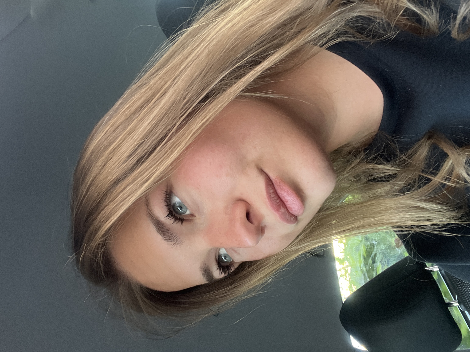A young woman is depicted in a selfie taken from inside a vehicle. The background suggests an outdoor environment, possibly viewed through the car window. The woman has light brown hair and light colored eyes. She is wearing a black top. Due to the lack of specific details in the image, it's impossible to determine the exact location in Budva, Montenegro.
