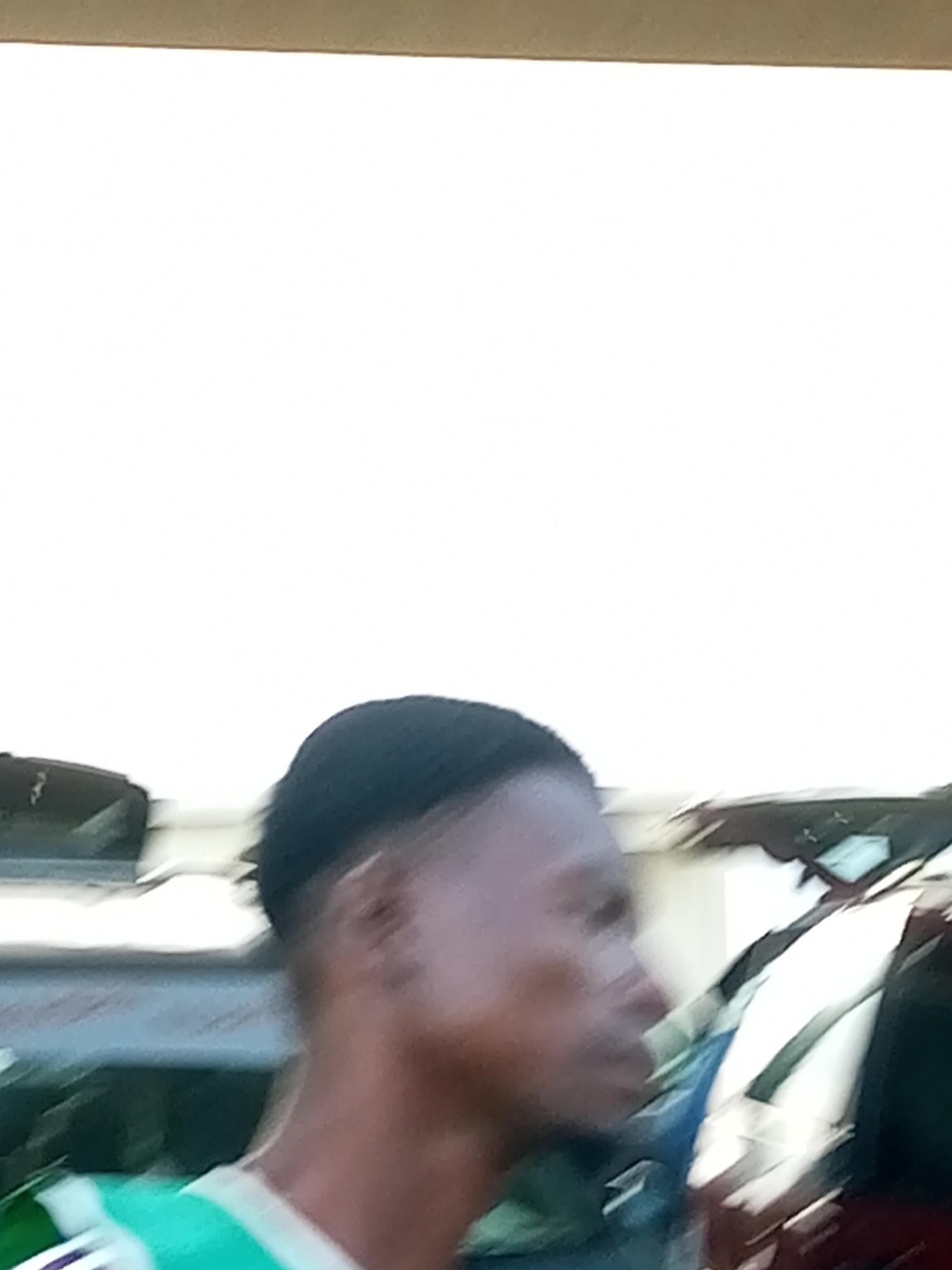 The image shows a close-up profile view of a young Black person with dark, short hair, wearing a green and white t-shirt. The person is looking to the left, with their head tilted slightly downwards. In the background, out of focus, are several vehicles, including what appears to be a dark-colored SUV or car and another vehicle that is partially visible. The overall setting appears to be outdoors, possibly a street or a parking lot, in the city of Gombe, Nigeria, given the context provided. The lighting suggests it is daytime.