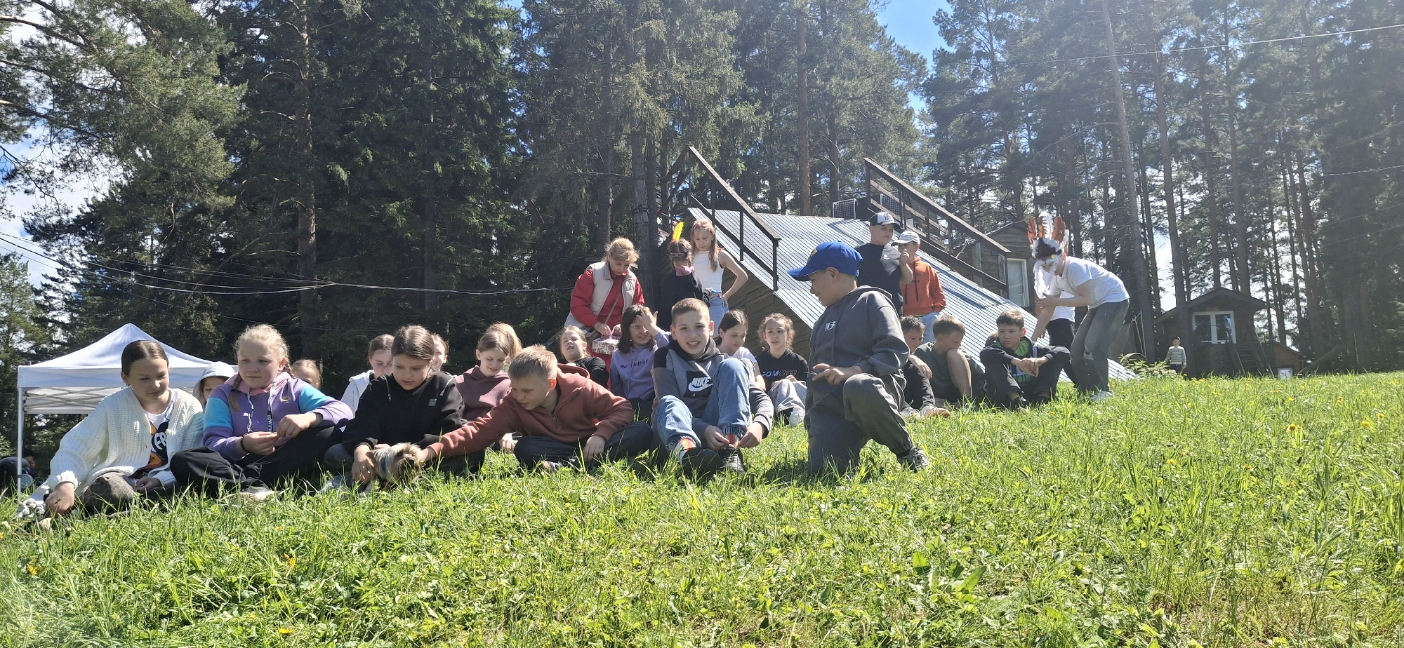 A group of children is sitting on a grassy hill in Прикамский, Russia. They are facing the camera. There is a building behind them, and trees in the background. Some of the children are interacting with pets.