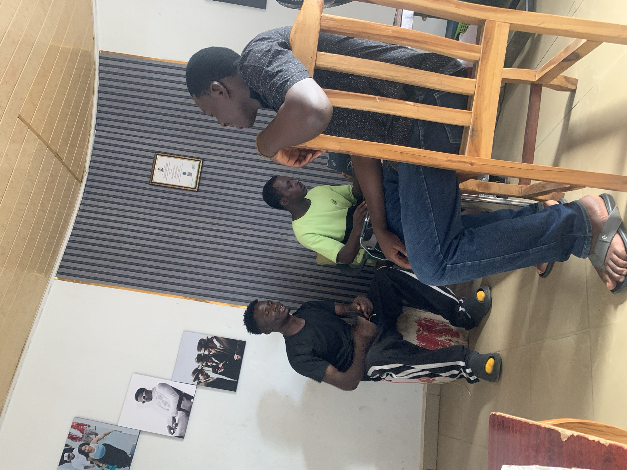 An indoor scene in Jos, Nigeria, captures three young men engaged in a casual gathering or conversation. The image is taken from a slightly elevated perspective within a room.
In the foreground, a man with short, dark hair is leaning forward over a light wooden chair. He wears a black patterned short-sleeved shirt, blue jeans, and grey flip-flops, and is looking downwards and to the left. His right arm rests casually on the chair's back.
To his left and slightly behind, a second man is seated on a low cushion or stool. He is dressed in a black short-sleeved t-shirt and black track pants with white stripes, and his feet are in black footwear with visible yellow soles. He looks slightly upwards and to the right, his mouth open as if speaking or reacting.
Further back, a third man is seated on a chair, wearing a neon green short-sleeved t-shirt. He also has short, dark hair and is looking towards the left, appearing attentive to the interaction.
The room features a white wall on the left adorned with four square photographic prints, depicting various subjects including a group of three men in hats and a man in white clothing and sunglasses. The wall in the background has horizontal grey and light-colored stripes, displaying a framed certificate or document with a gold border, possibly bearing a Nigerian coat of arms (text is illegible). A wooden panel ceiling or upper wall section is visible on the far left. The floor is tiled in a light color, and a small red mark is visible on the floor near the seated man in track pants. The lighting suggests well-lit indoor conditions.