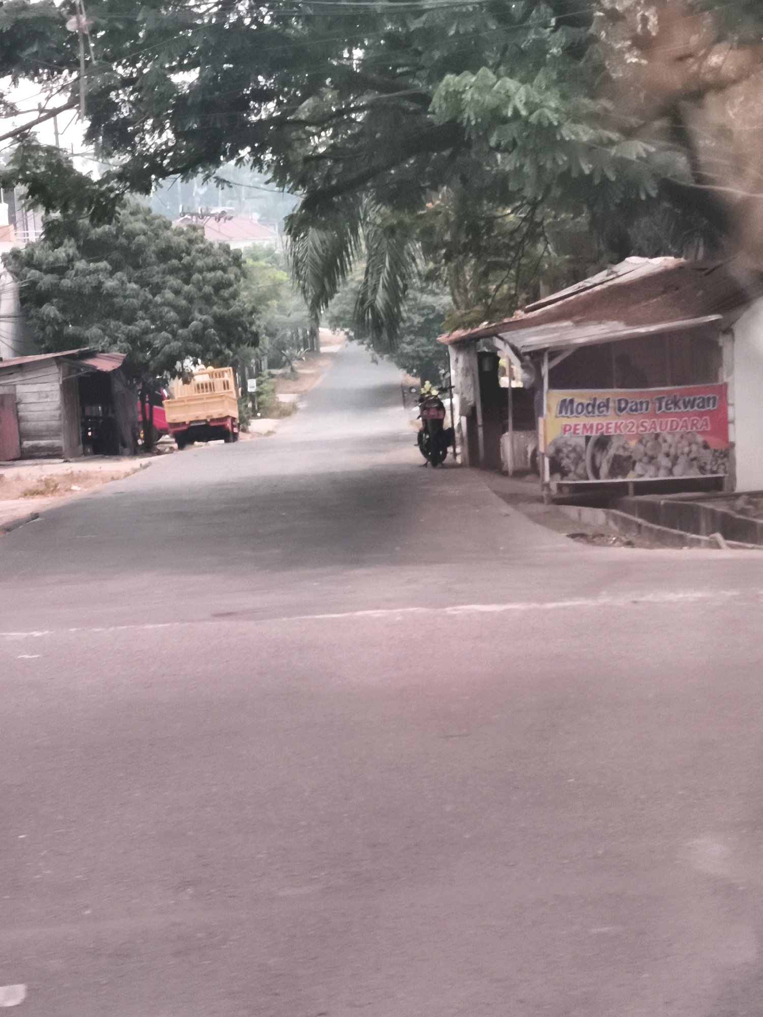 The image depicts a street scene in Palembang, Indonesia, likely in a more rural or suburban area given the presence of trees, a simple roadside stall, and a yellow truck. On the right side of the road, a small building with a rusted corrugated metal roof serves as a roadside food stall. A banner advertising "Model Dan Tekwan PEMPEK 2 SAUDARA" is prominently displayed on the front of the stall, indicating it sells local Palembang delicacies like Tekwan and Pempek. Behind the banner, an image of Pempek is visible. A motorcycle with a rider is parked near the stall. To the left of the stall, a yellow truck is parked on the side of the road, with a small wooden structure also visible. The road is paved and appears to be a two-lane street, curving uphill and surrounded by lush greenery, including large trees and palm fronds hanging over the road. The lighting suggests it could be overcast or early morning/late afternoon. The overall impression is a snapshot of everyday life in a Palembang neighborhood, with commerce and transportation coexisting along a tree-lined street.