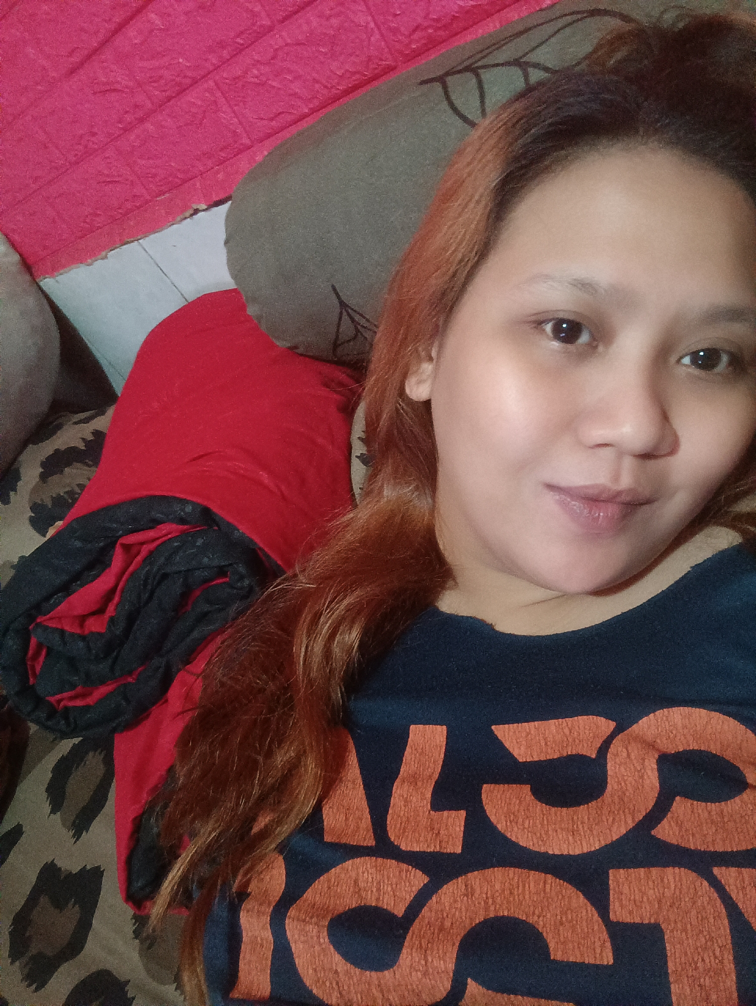 In an indoor setting, likely a bedroom in Depok, Indonesia, a woman is captured in a selfie. She has light brown hair, which appears to be dyed with reddish-brown highlights. Her face is the central focus, with her eyes looking directly at the camera, and a slight smile on her lips. She is wearing a dark blue t-shirt with large, orange, block-style letters that are partially visible.
The background of the image reveals a variety of textures and colors. To the left, a textured, bright pink surface is visible, possibly a decorative wall panel or cushion. Below this, a red fabric, appearing to be a rolled blanket or throw, is draped over a patterned surface that resembles leopard print. Further to the right, a grey pillow or cushion is visible. The overall impression is a casual and personal space.