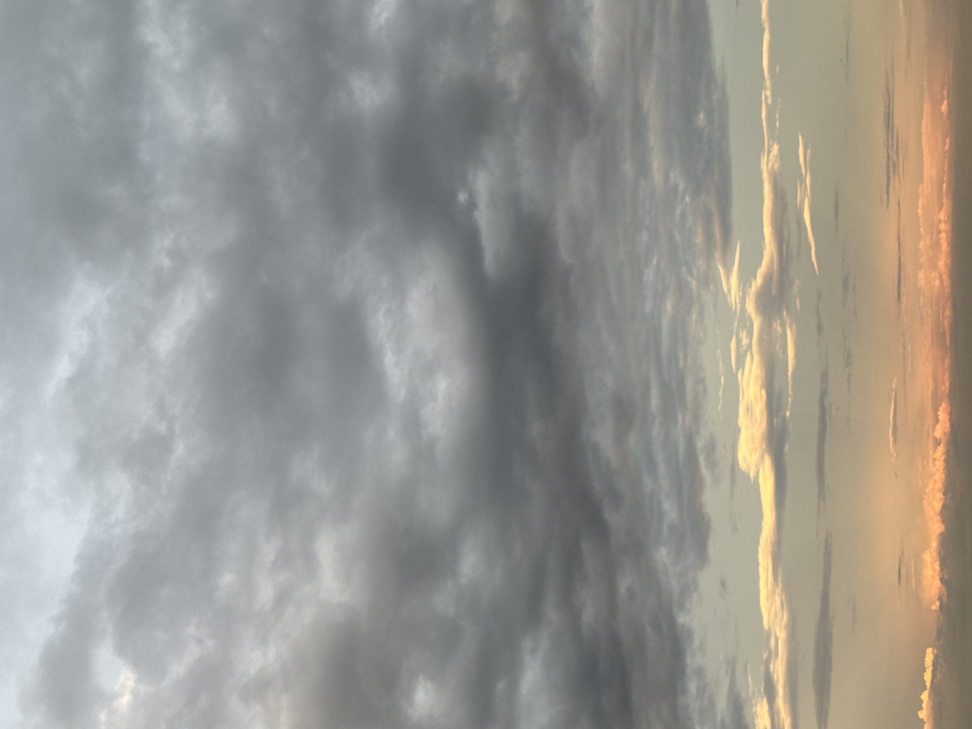 The image depicts a sky filled with clouds. The left side of the image shows predominantly gray, dense clouds, suggesting an overcast or potentially stormy weather. Towards the right side, the clouds transition to lighter shades of gray and white, with hints of orange and yellow light peeking through, possibly indicating sunrise or sunset. The photo is taken from a low angle. No people, subjects, or objects are visible besides the sky. The location and actions are impossible to determine. The city of Dikaya Gar', Russia, is not evident in the picture.
