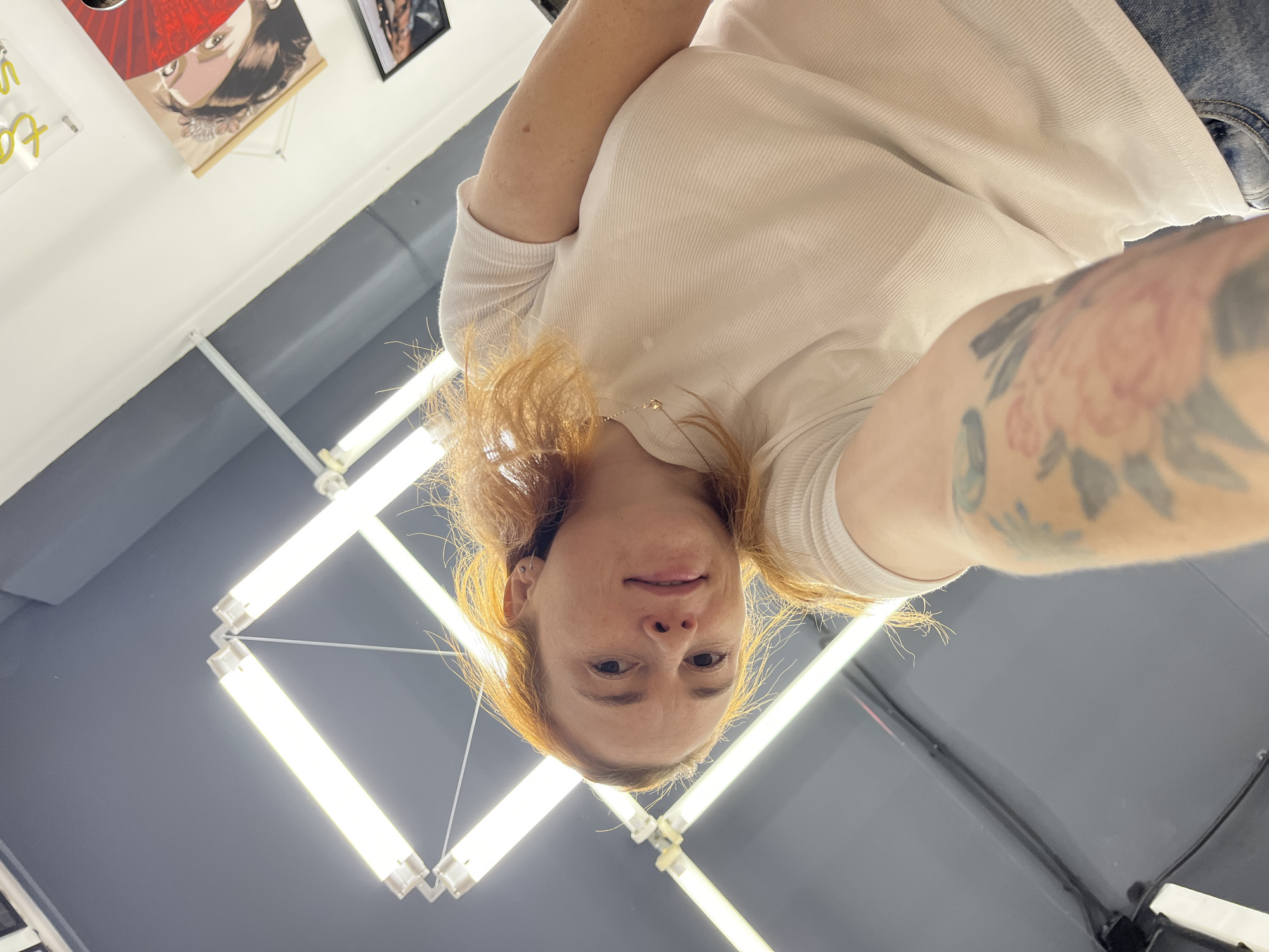 In a dimly lit room with grey walls and ceiling, a woman with red hair and fair skin, wearing a white t-shirt and blue jeans, is taking a selfie. Her left arm is extended towards the camera, revealing a tattoo of a pink flower on her forearm. She has a faint smile on her face, and her hair is slightly messy. The room appears to be a studio, possibly a tattoo parlor, indicated by the neon sign and framed artwork on the wall. Fluorescent lights are mounted on the ceiling, casting a bright, even illumination. The overall impression is that of a candid moment captured in a creative space. The location is identified as Chaikovsky, Russia.