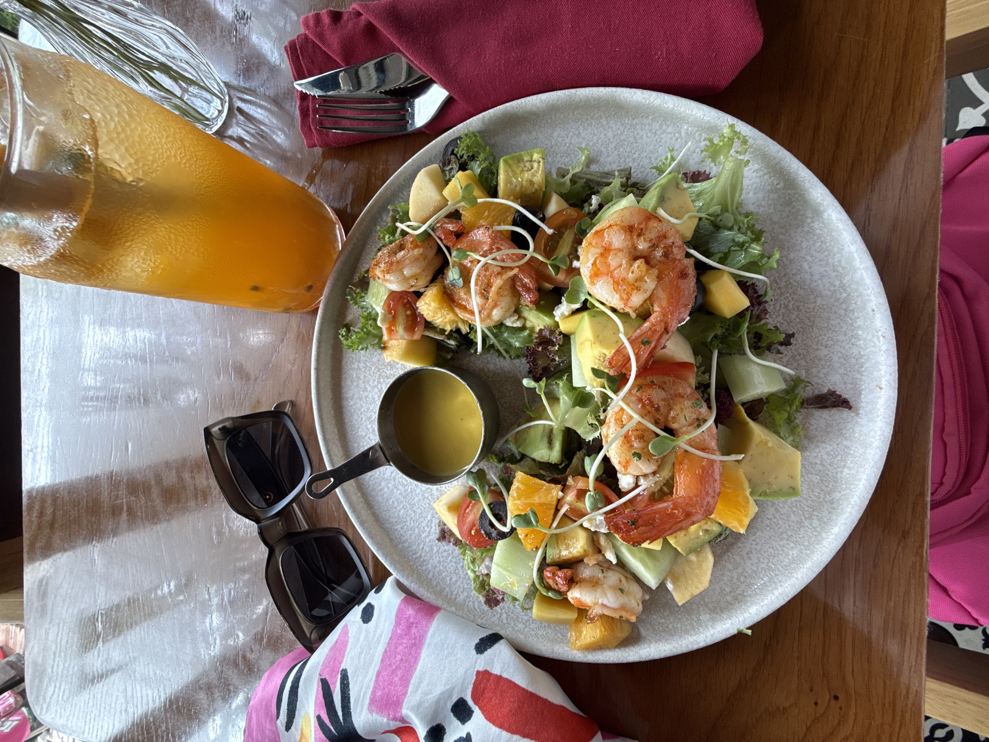 The image is a top-down shot of a table arrangement. A shrimp salad is the main focus, presented on a speckled off-white plate. The salad comprises lettuce, avocado, mango, orange slices, cherry tomatoes, olives, sprouts, and grilled shrimp. A small metal container, filled with a yellow sauce, is placed on the side of the plate.
Next to the plate, to the left, lies a pair of dark-rimmed sunglasses and a colorful patterned scarf. A glass pitcher, filled with orange liquid, is positioned at the top left. Adjacent to the pitcher is a maroon napkin with cutlery (knife and fork) on top.
The table has a wooden surface. In the background, portions of a pink garment and a black and white patterned chair are visible.
Given the style of cuisine and the tropical ingredients, it's plausible that the location is in Nha Trang, Vietnam, a coastal city known for its seafood. The scene suggests a casual dining setting, likely an outdoor cafe or restaurant. There are no people directly visible in the image.