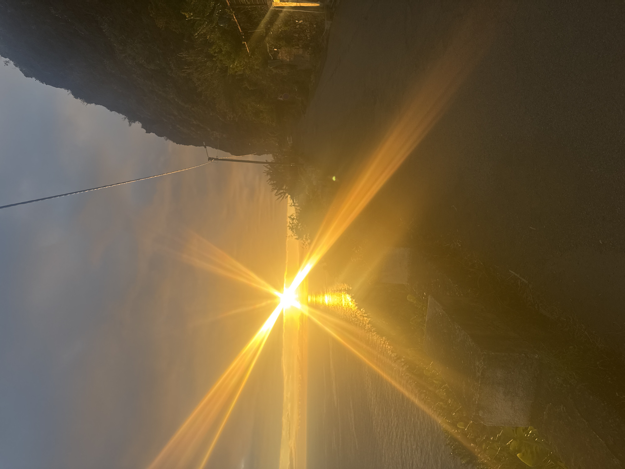 The image depicts a street scene in Ponta do Sol, Portugal, at sunset. The sun's intense glare dominates the center of the frame, reflected off the water and obscuring details. The road extends to the right, with a stone wall separating it from the water. The mountain line rises on the left. A power line stretches across the top left of the frame. Vegetation lines the road, and the scene is bathed in the golden light of the setting sun.
