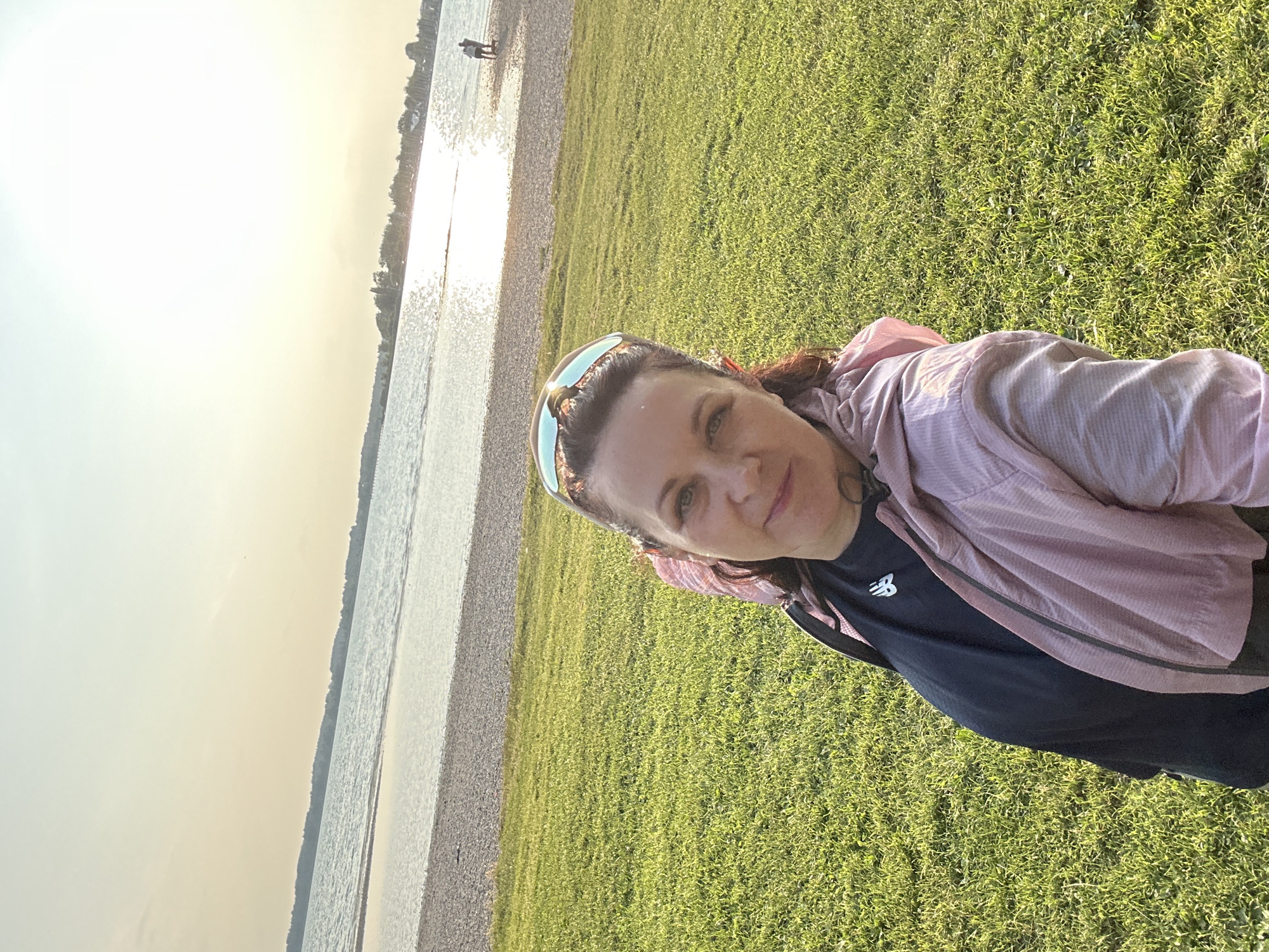 In Chieming, Germany, a woman is captured in a selfie by the Chiemsee lake during sunset. She is positioned in the foreground, with her upper body angled towards the camera. She has dark hair pulled back and is wearing sunglasses on her head. She is dressed in a black t-shirt with a white logo and a light pink hooded jacket. The background reveals the Chiemsee lake, its waters reflecting the golden hues of the setting sun. Along the shore, there is a gravel beach, and further in the distance, a treeline and buildings are visible. Two figures are seen walking on the beach in the middle ground. The foreground is dominated by a lush green lawn. The lighting suggests it is late afternoon or early evening, with warm, soft sunlight illuminating the scene.