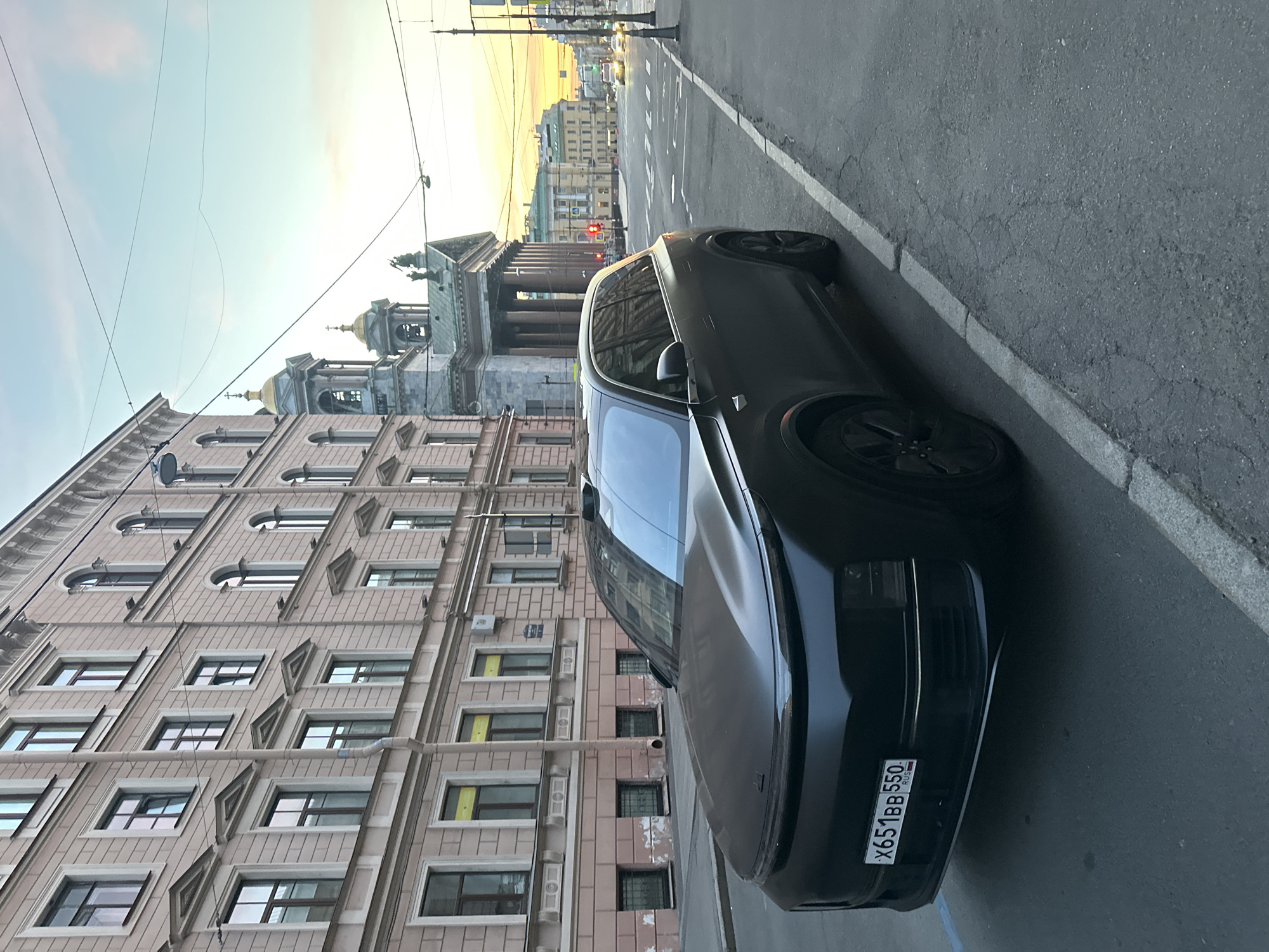 A matte black SUV is parked on the side of a street in front of a multi-story building with a light pink facade and numerous windows. The car has a license plate that reads "X651BB 550 RUS". In the background, across the street, there's a large domed building with a green roof and a bell tower, and further down the street, several other buildings and traffic lights are visible. The sky is a pale blue with hints of orange and yellow from what appears to be a sunset. The street is paved and cracked in places, with a sidewalk separating it from the building. There are overhead wires visible in the sky. No people are clearly visible in the image.