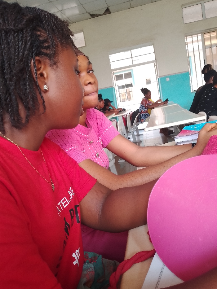 The image shows two young women seated at desks in a classroom or waiting area. The woman in the foreground is wearing a red t-shirt and has braided hair. The woman behind her is wearing a pink top. Other individuals are seated at desks in the background. The location is OGBOKO, Nigeria. The environment appears to be a public space with light blue walls and metal window frames.