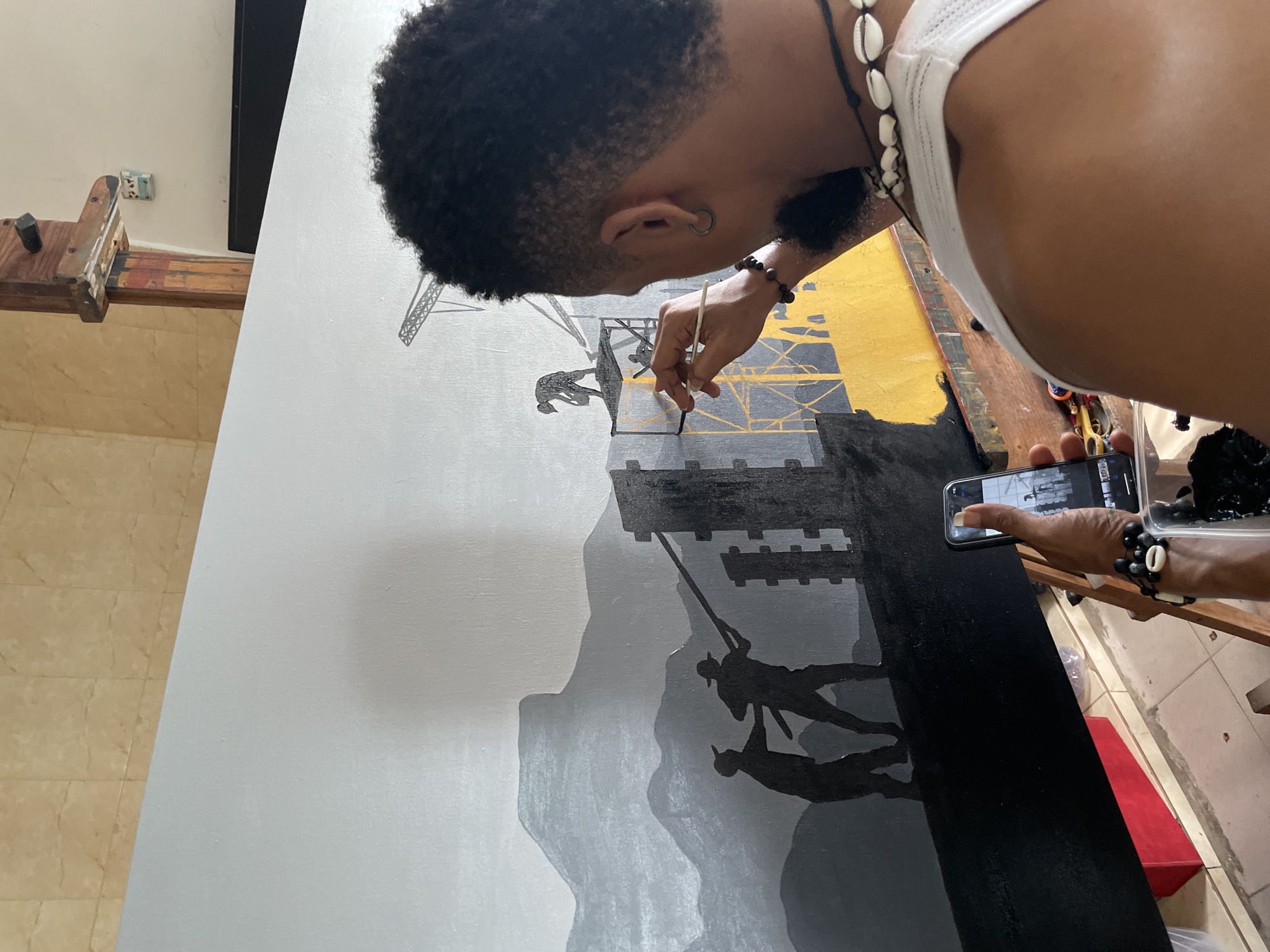 A person is painting on a large canvas, possibly in Ado, Nigeria. The painting depicts silhouetted figures climbing or swinging from a structure amidst a backdrop of mountains or cliffs. The painter, wearing a white tank top and necklaces, holds a brush in one hand and a phone in the other, appearing to photograph the painting. The canvas is positioned on an easel. The floor is tiled. A second canvas is on the right of the painter.
