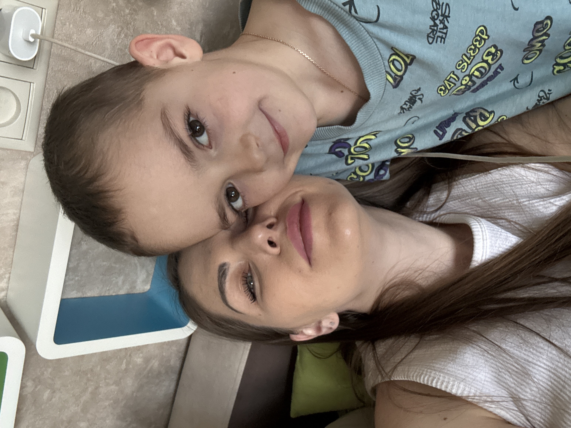 A woman and a young boy are lying closely together, face-to-face. The boy, on top, is wearing a light blue t-shirt with yellow and purple skateboard-themed cartoon images. He is also wearing a thin gold chain around his neck. The woman has long brown hair and is wearing a white shirt. They are lying on a surface that appears to be a sofa or bed with colorful geometric shapes and patterns visible on a nearby object. The location is likely indoors, but further details to confirm the location in Раздолье, Russia, are absent.