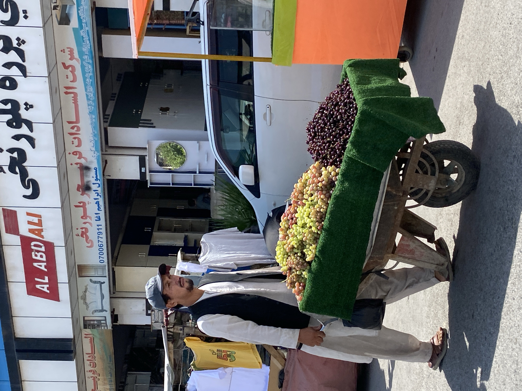 In Kandahar, Afghanistan, a man wearing a gray baseball cap, a dark vest, and a light-colored tunic and trousers pushes a fruit cart on a paved street. The cart is loaded with two varieties of grapes: a large pile of green and red grapes and a smaller pile of dark purple grapes. The cart is covered with a dark green cloth. In the background, a white van is parked and behind it, a store facade with signs in both English and Pashto is visible. Clothing items are displayed on a rack to the left of the man. The sun casts sharp shadows on the ground.